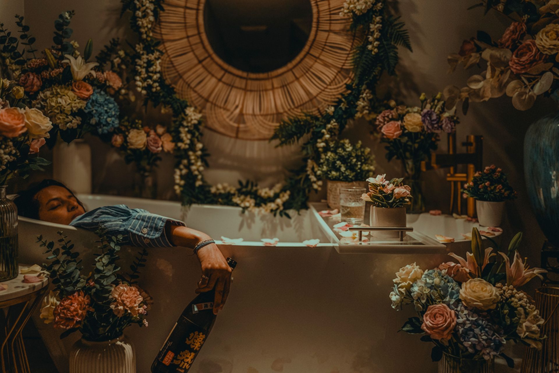 A person relaxes in a bathtub surrounded by abundant floral arrangements and greenery in a dimly lit, moody room.