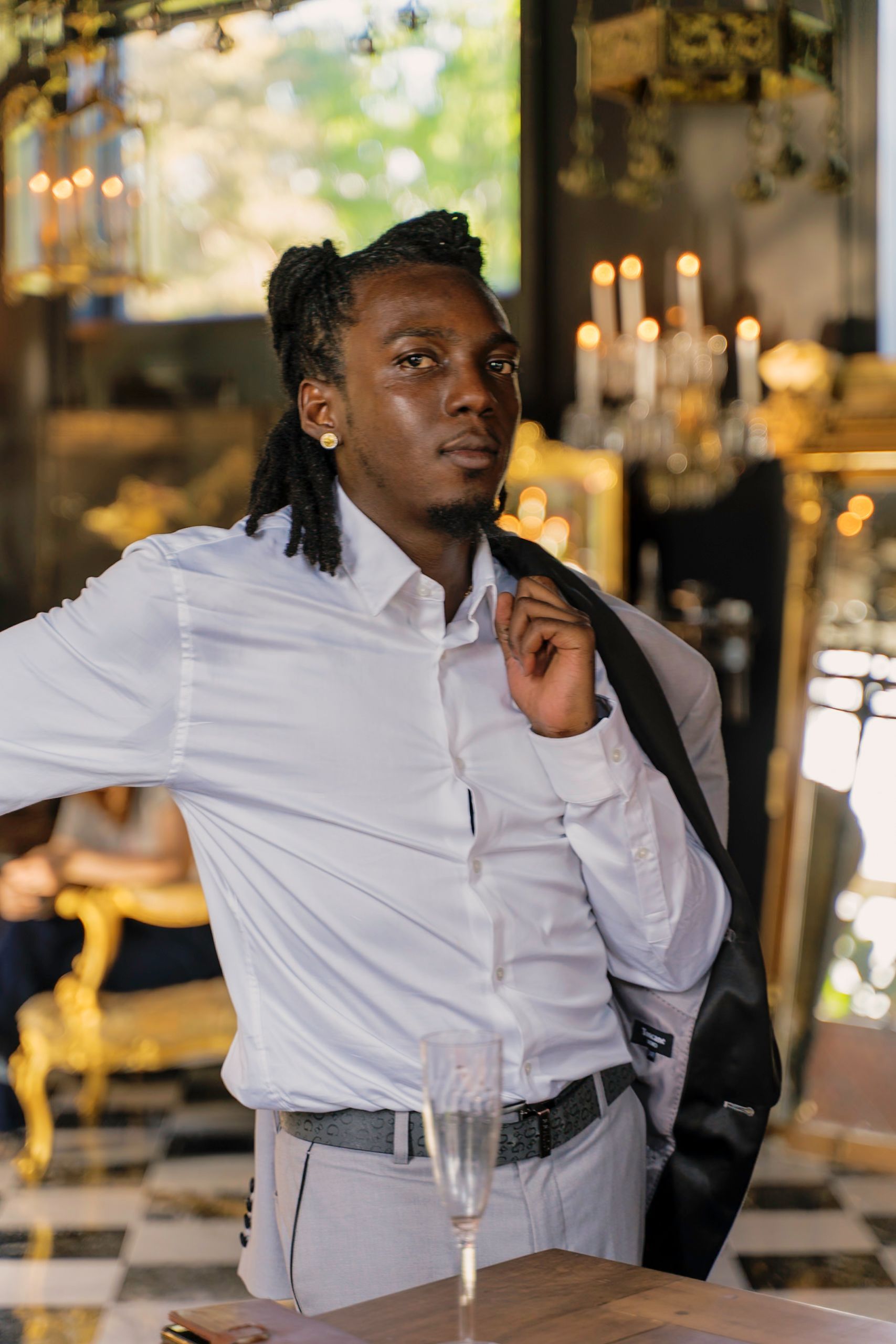 Man in white shirt adjusts his blazer, standing in a luxury setting, with a champagne glass.