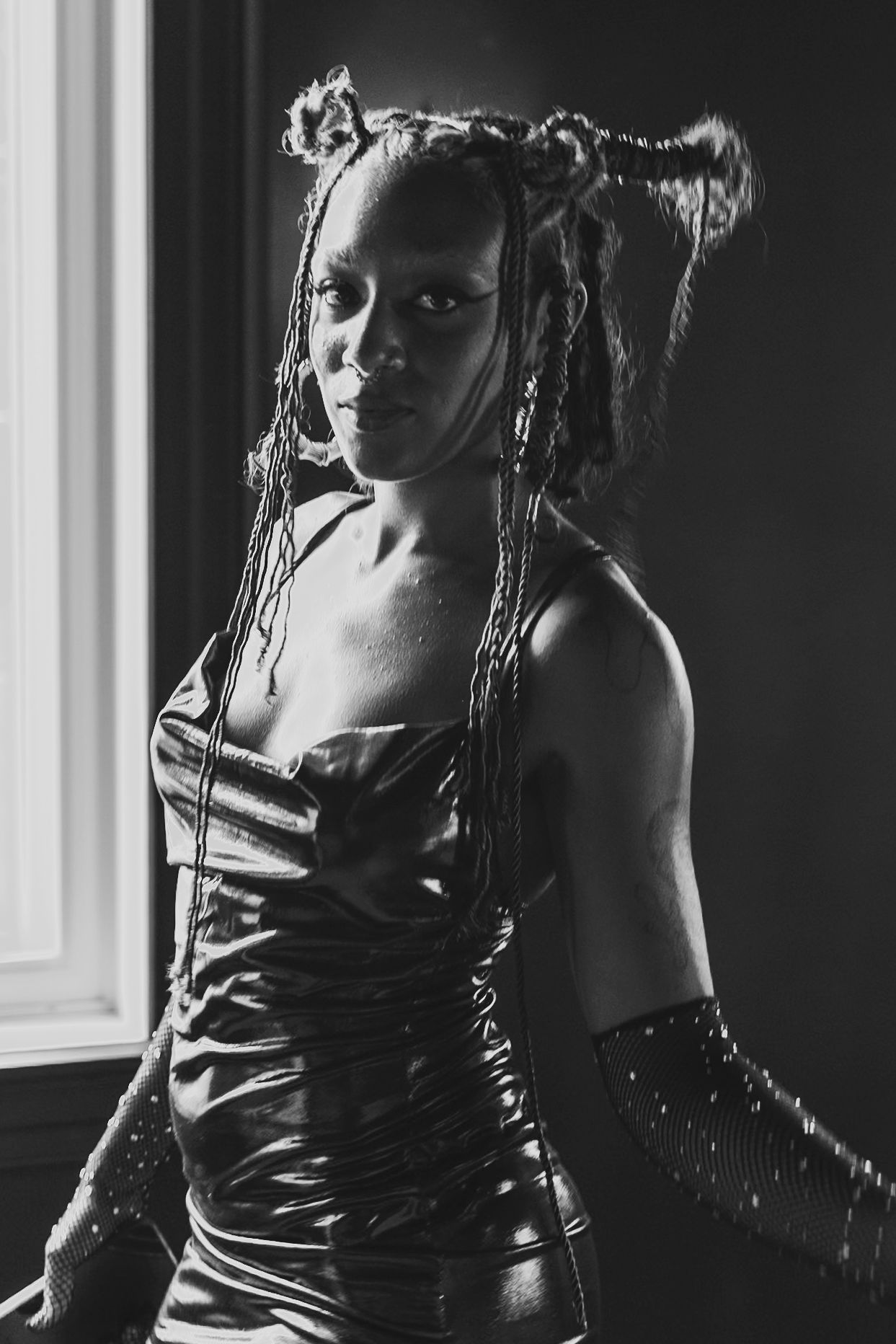 Woman in dark dress, poses near a window. Braided hair up, arms in gloves, looking towards the camera.