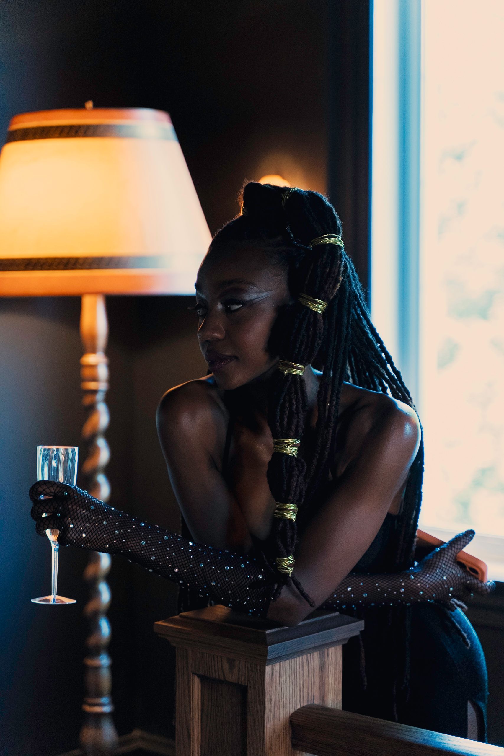 Woman with braided hair, holding champagne, leaning on a wooden banister near a window. Dimly lit room.