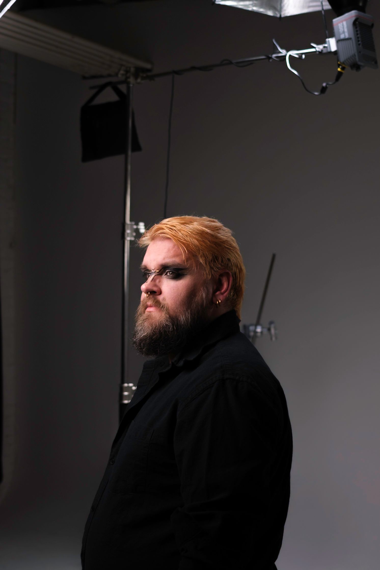 Man with orange-blonde hair and beard, wearing black coat, lit in a studio setting.