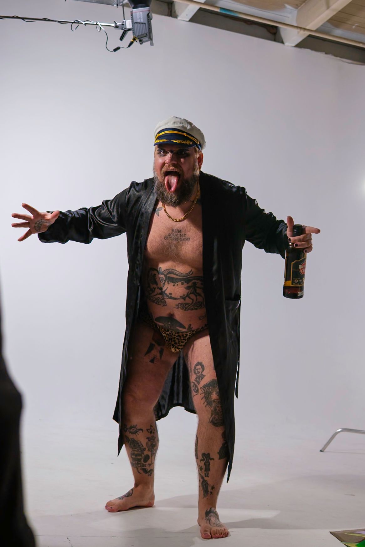 Man with tattoos, captain's hat, and open coat sticking out tongue, holding a drink, standing in a studio with white background.