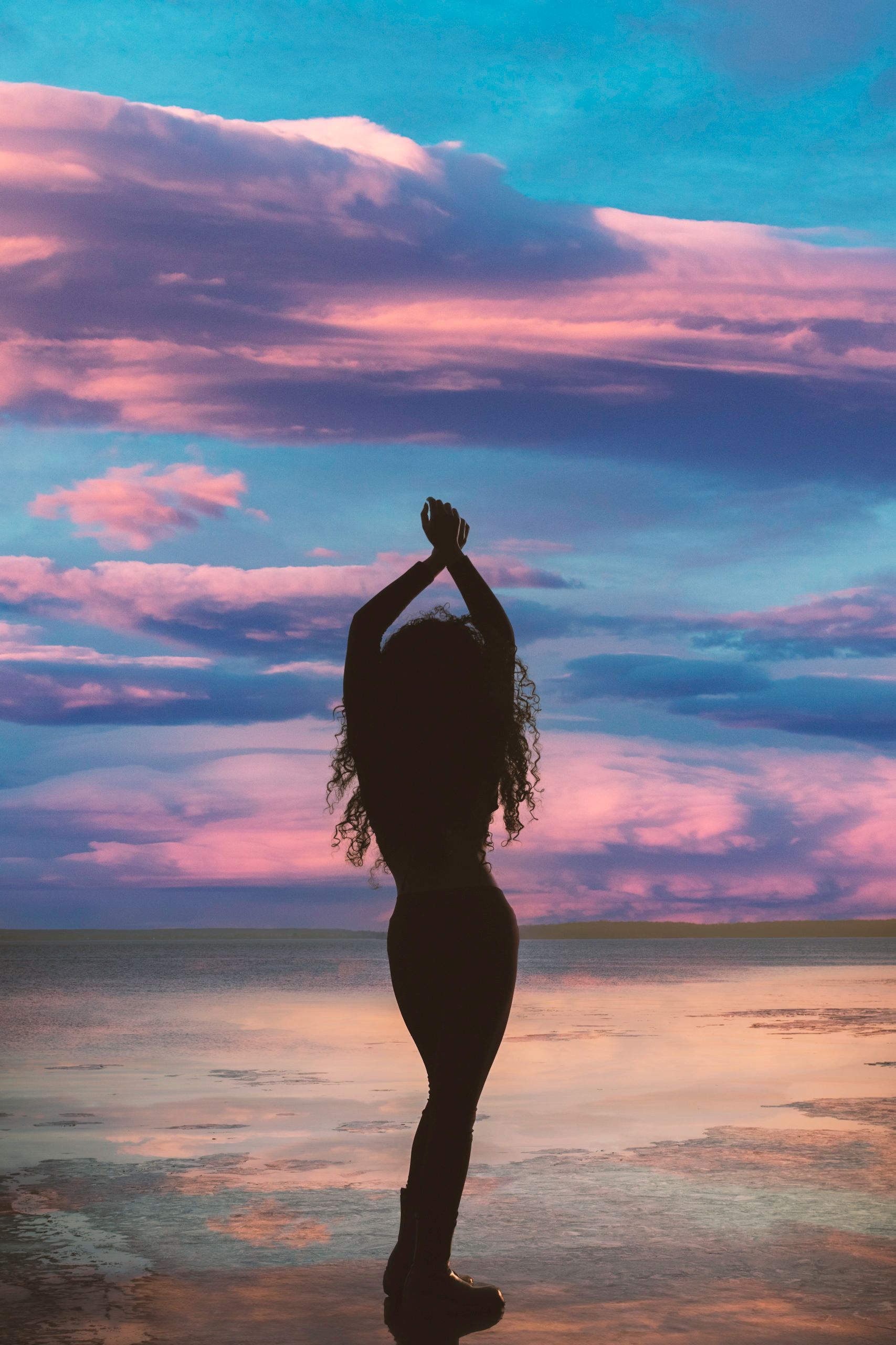 Silhouette of person with arms raised, standing on reflective surface. Sunset with blue, pink, and purple sky.