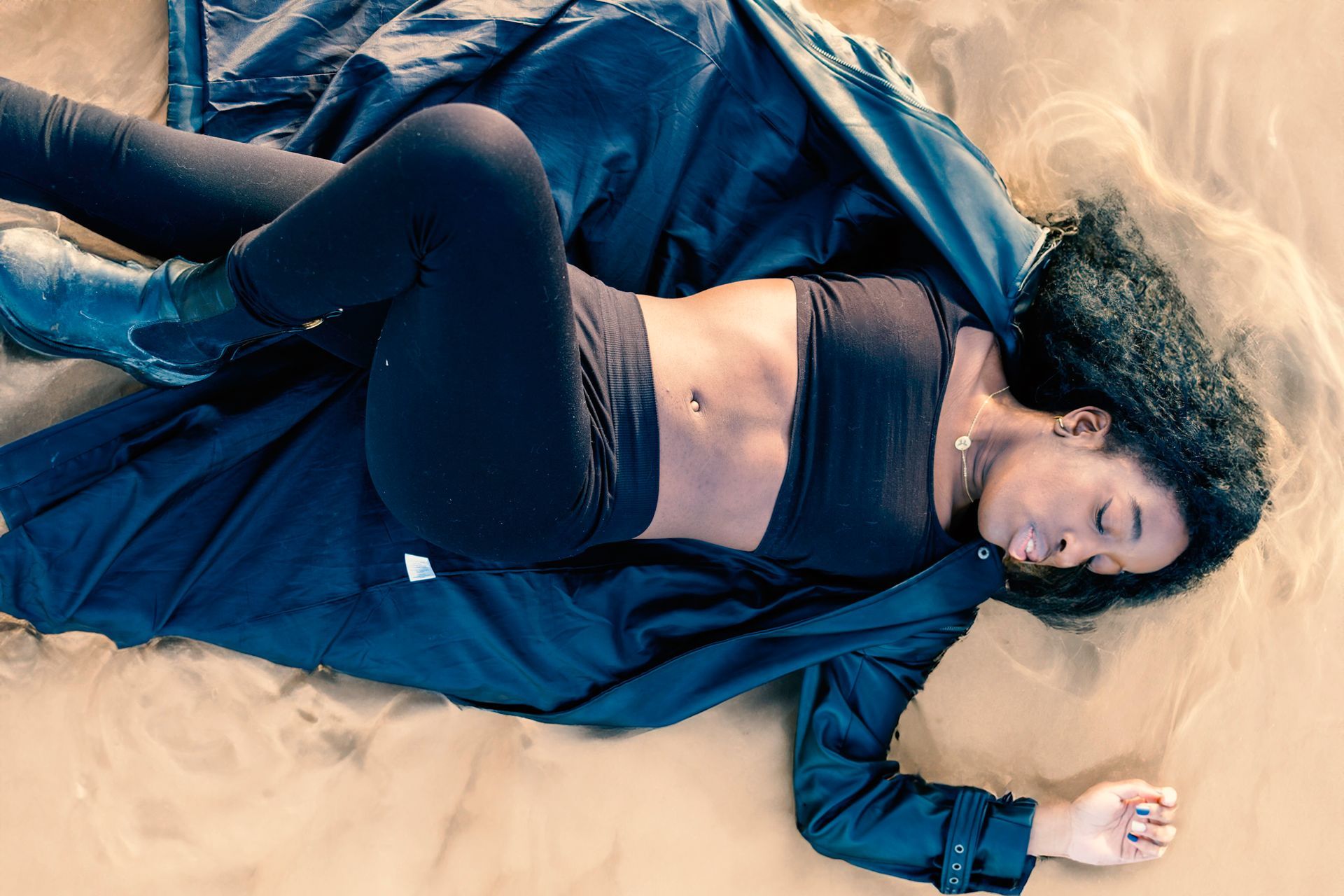 Woman in black top, leggings, and coat lies in sand, eyes closed.