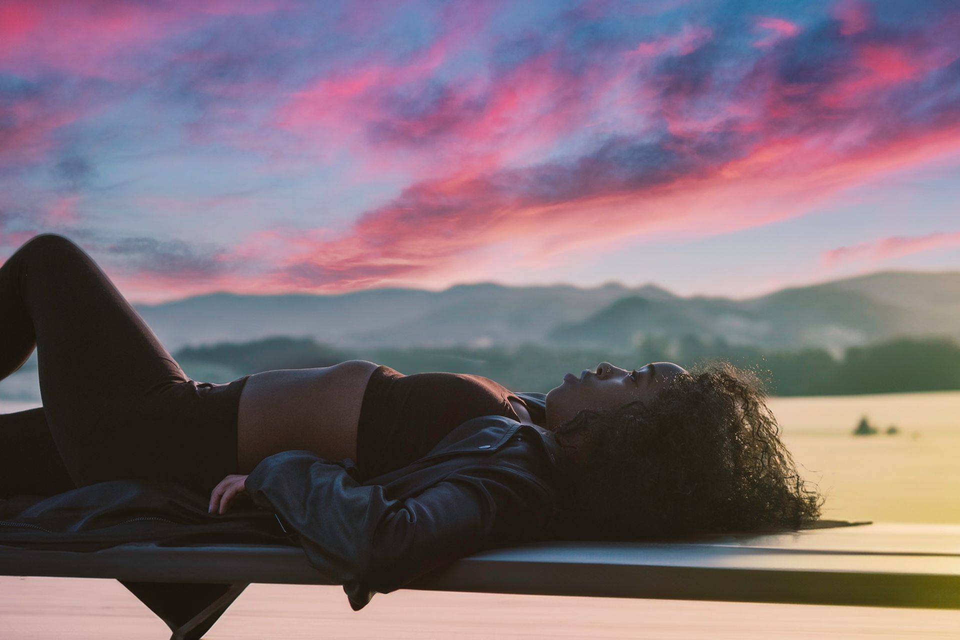 Woman reclines on a bench, gazing at a sunset over mountains and a lake.