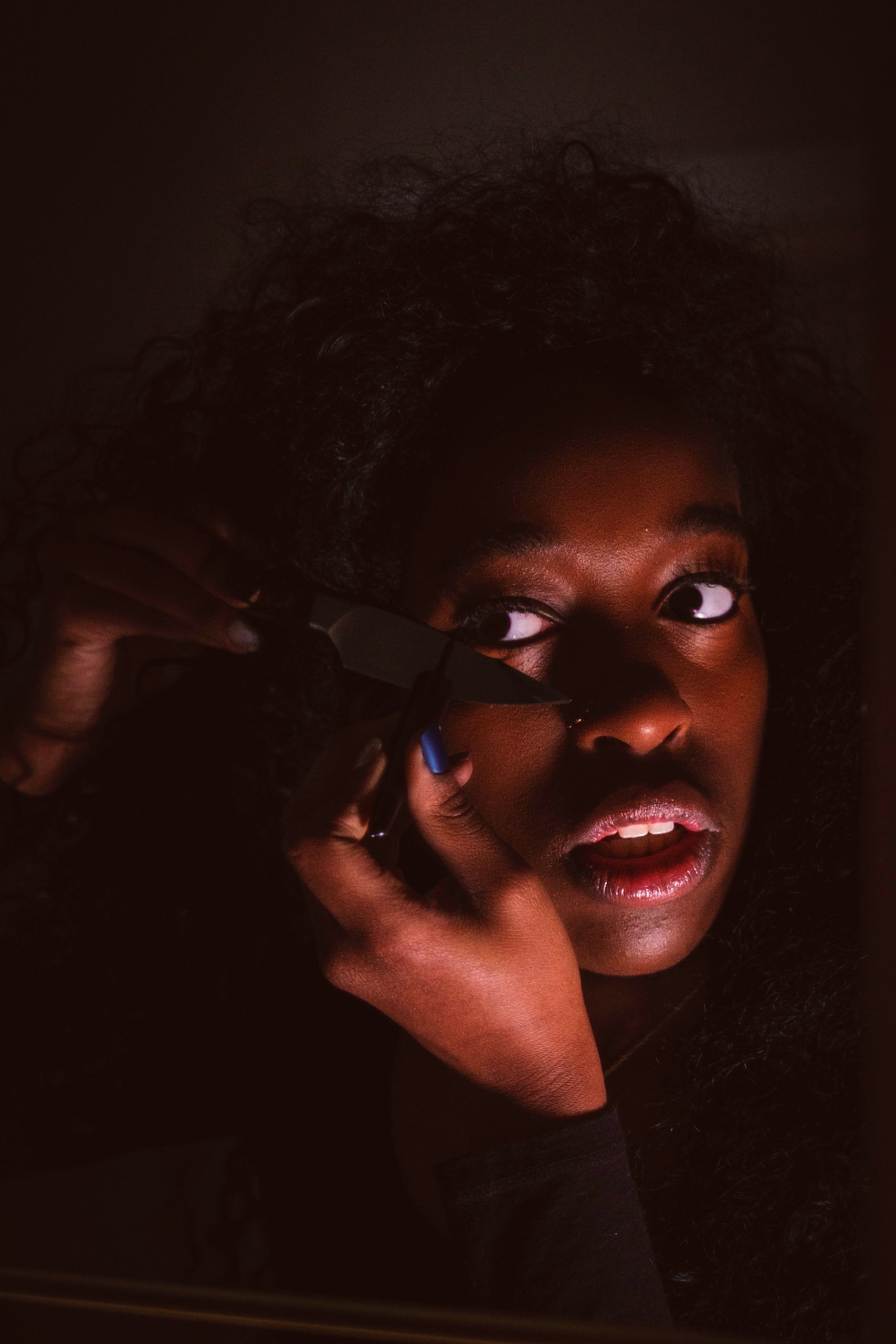 Woman applying makeup in a dimly lit setting. She looks slightly surprised, holding a makeup tool near her eye.
