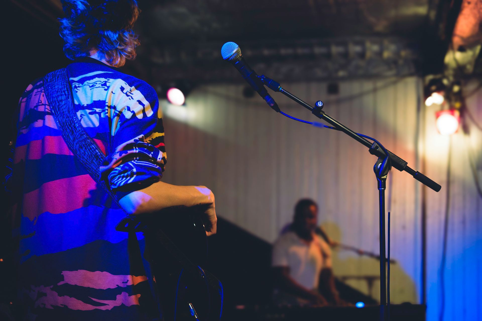 A person performing on stage, wearing a colorful shirt; microphone in front. Another musician in background.