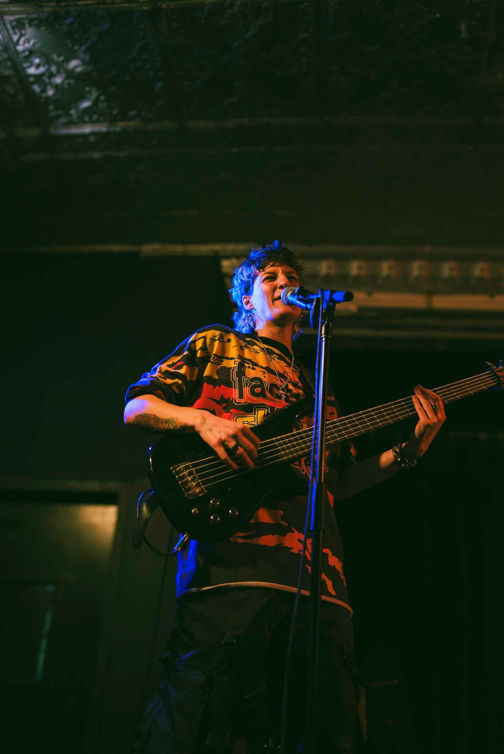 Person playing bass guitar and singing into a microphone onstage under colored lights.