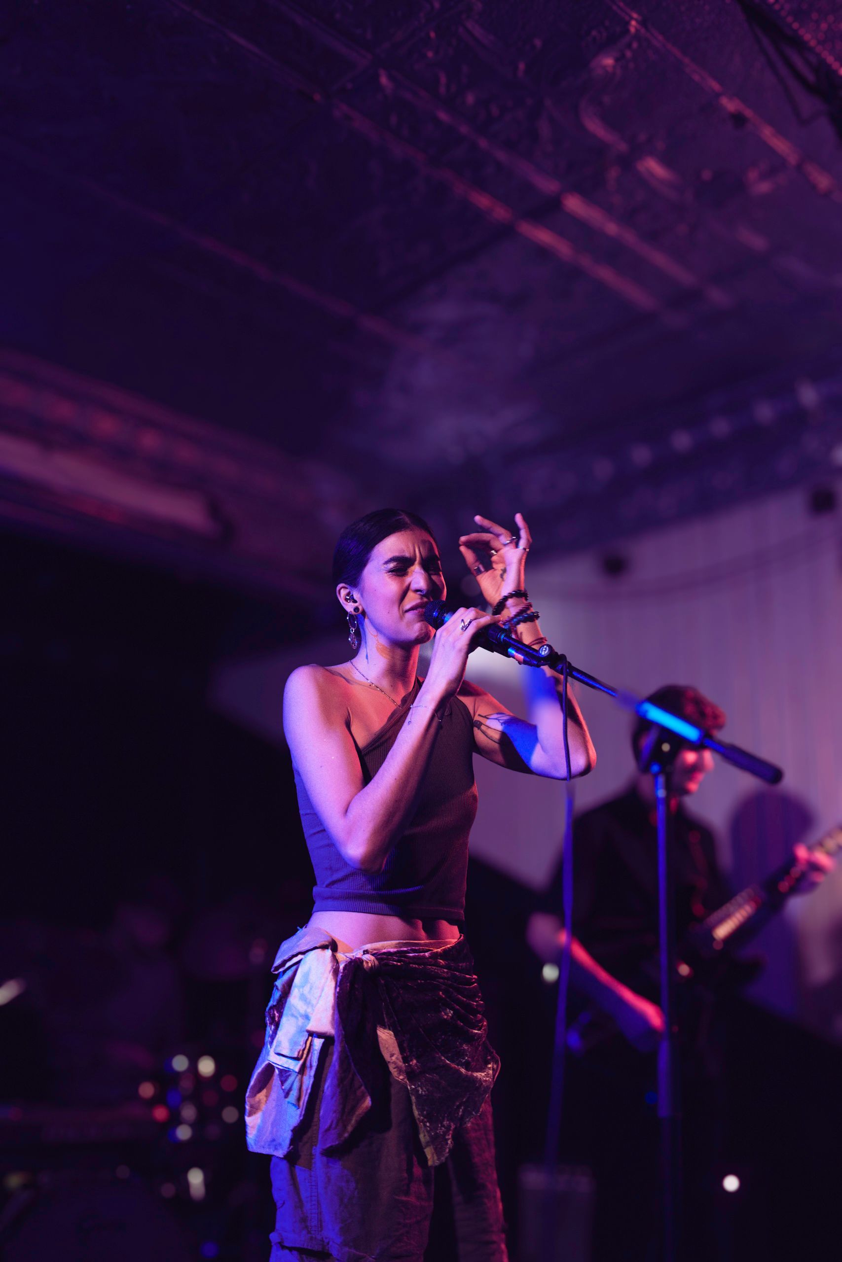 Woman singing into a microphone on stage, lit with purple and white lights. Another musician in background.