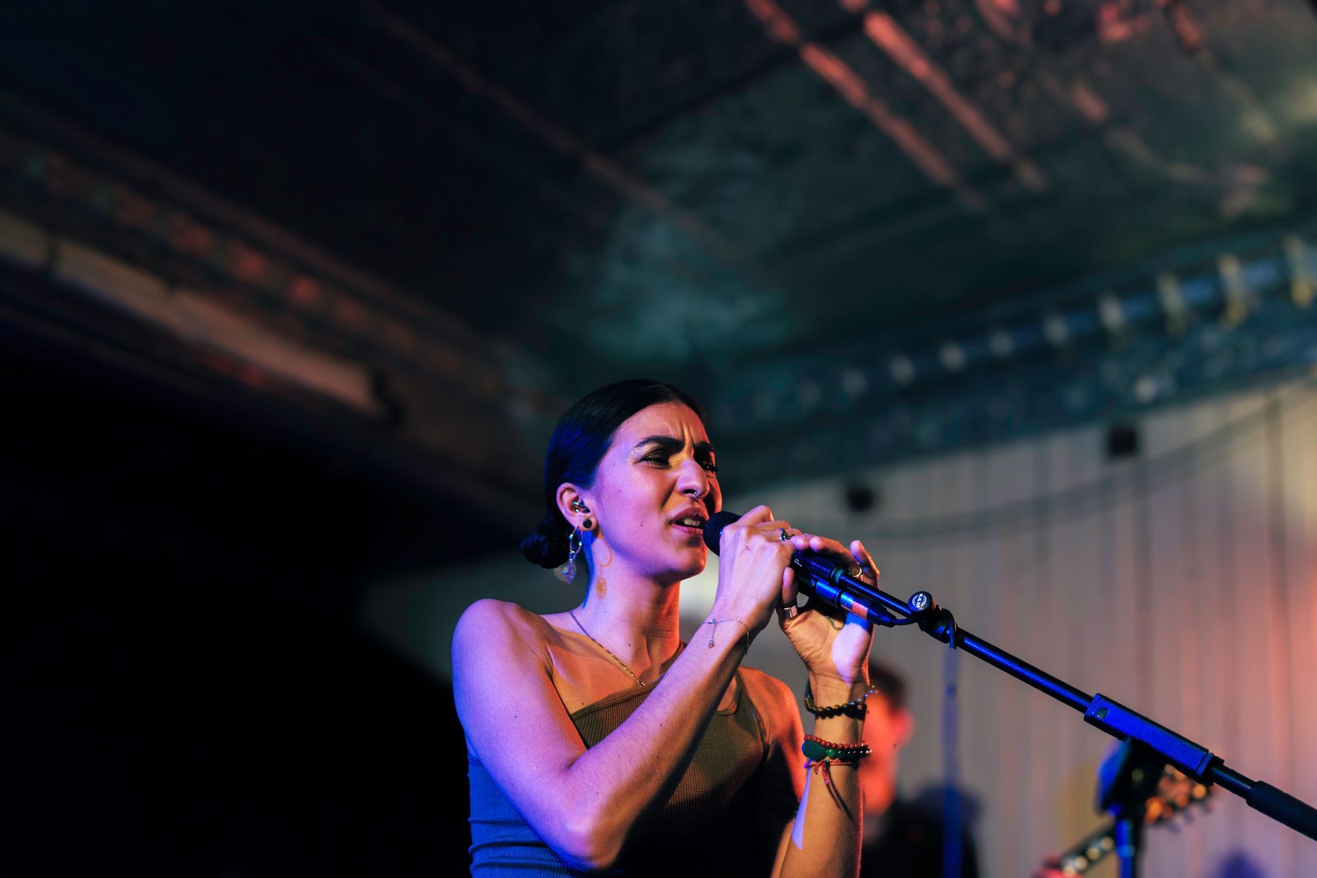 Woman singing into a microphone on stage, lit with blue and orange lights.