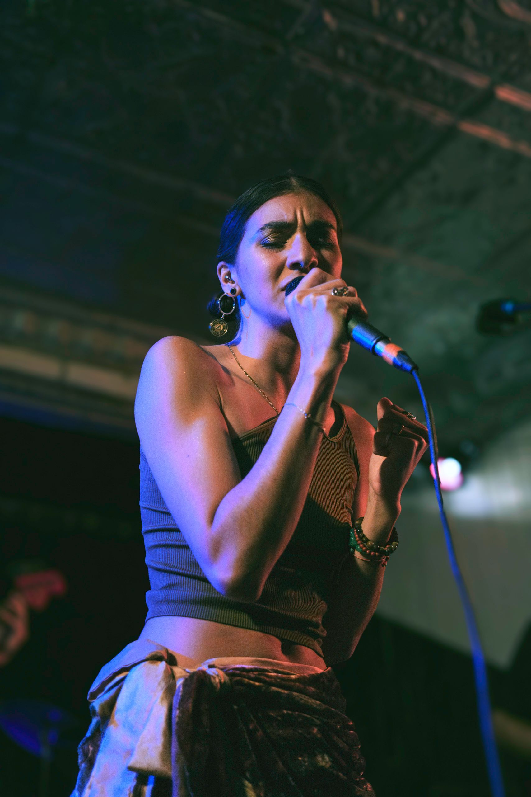Woman singing into a microphone onstage; wearing a patterned top and skirt. Moody lighting.
