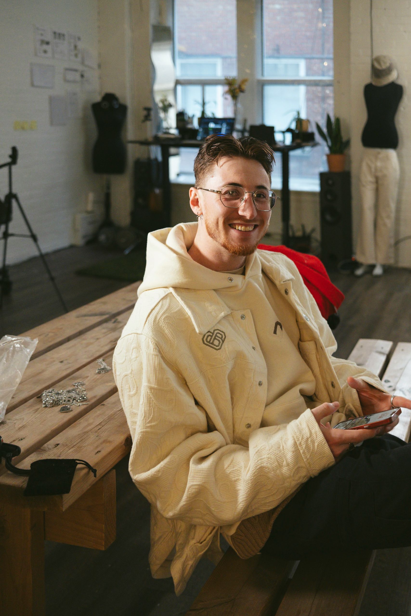 Man smiling, wearing a cream jacket, holding objects, sitting at a wooden table in a studio with sewing forms.