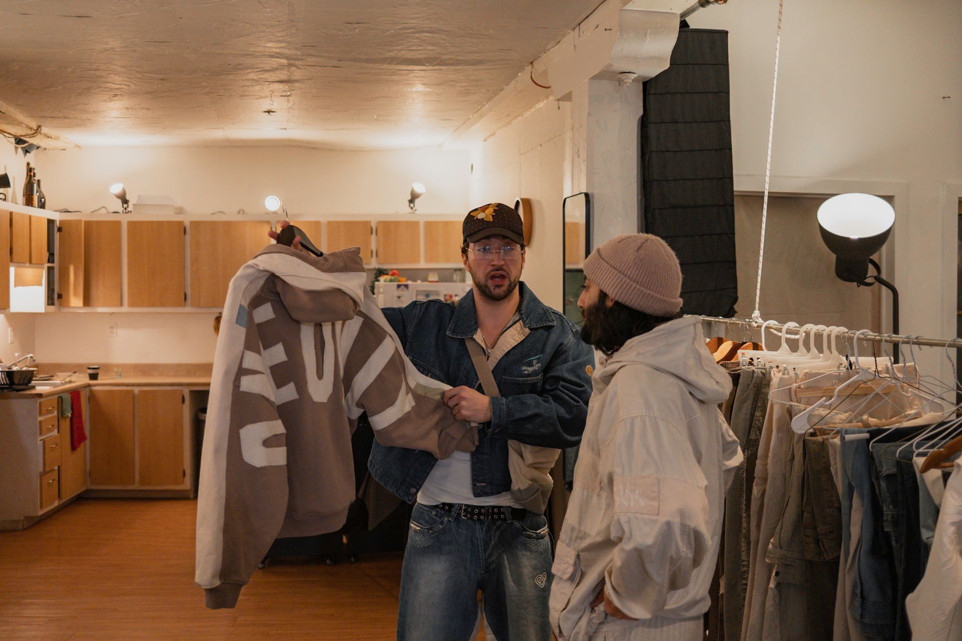 Two people in a shop, one holding a tan sweatshirt with large lettering, clothing rack in background.