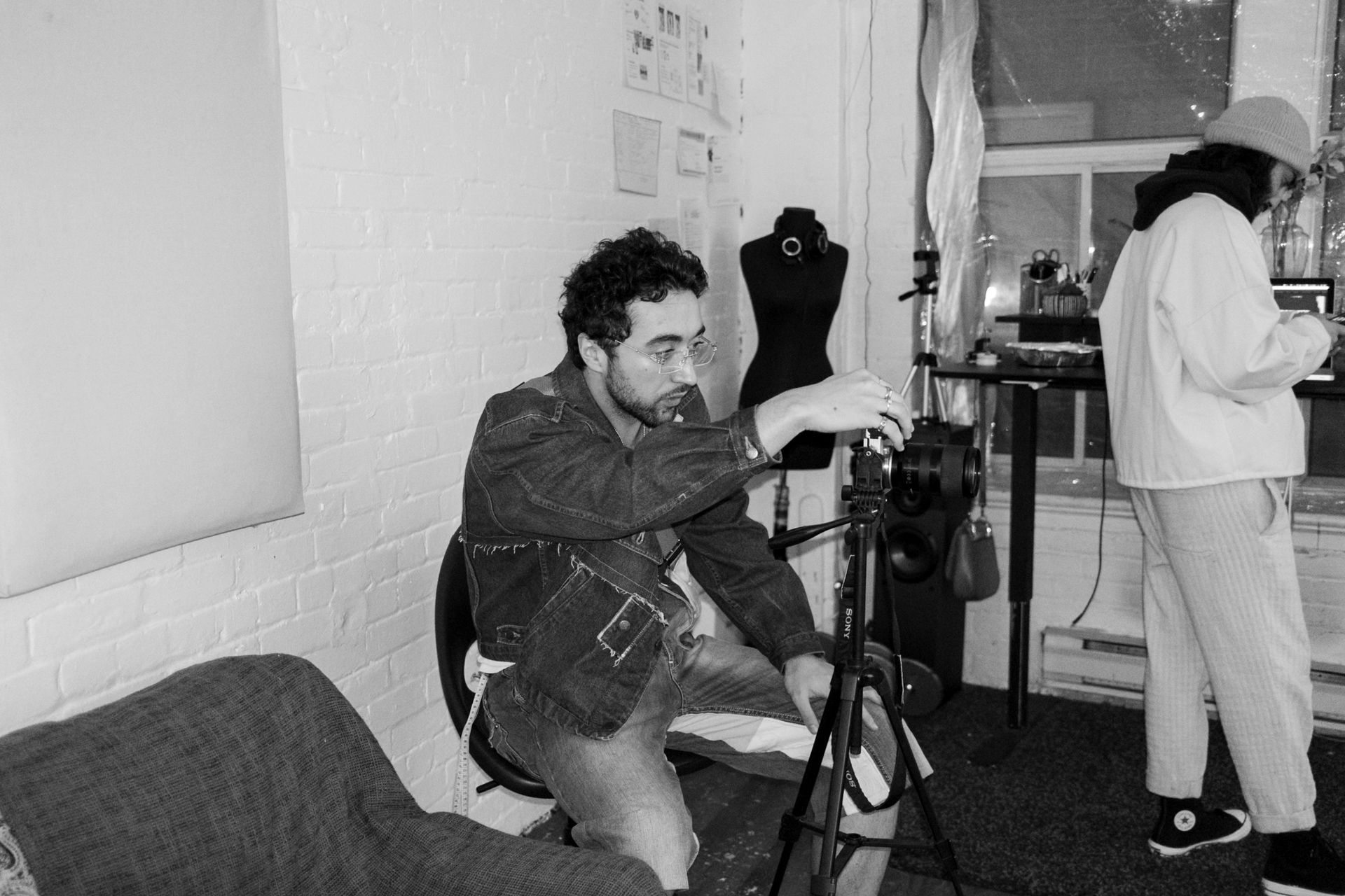 Man with camera on tripod, adjusting it. Another person in background. Indoor setting, black and white.
