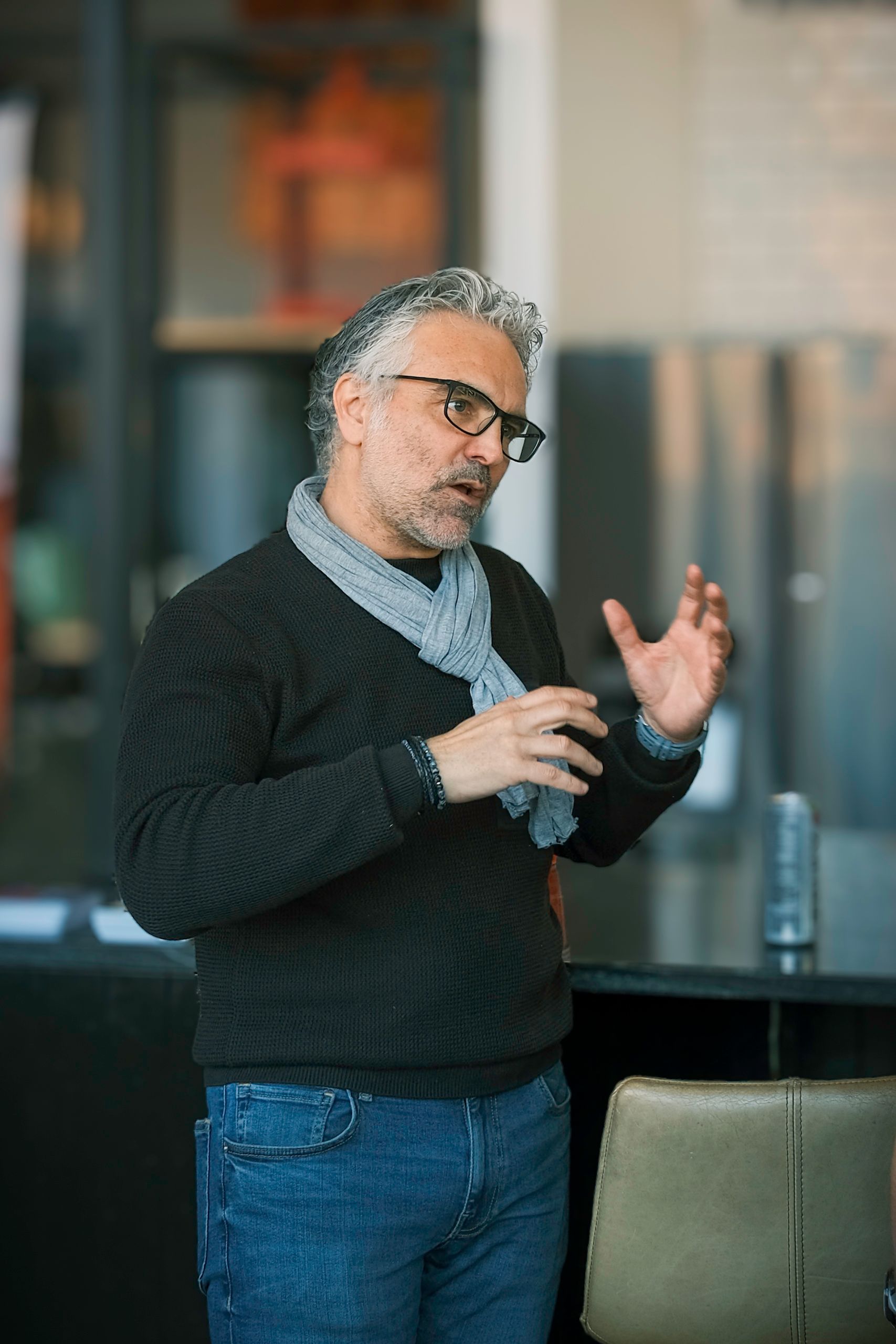 Man gesturing with hands, wearing glasses, black sweater, blue scarf, and jeans, speaking in a modern setting.