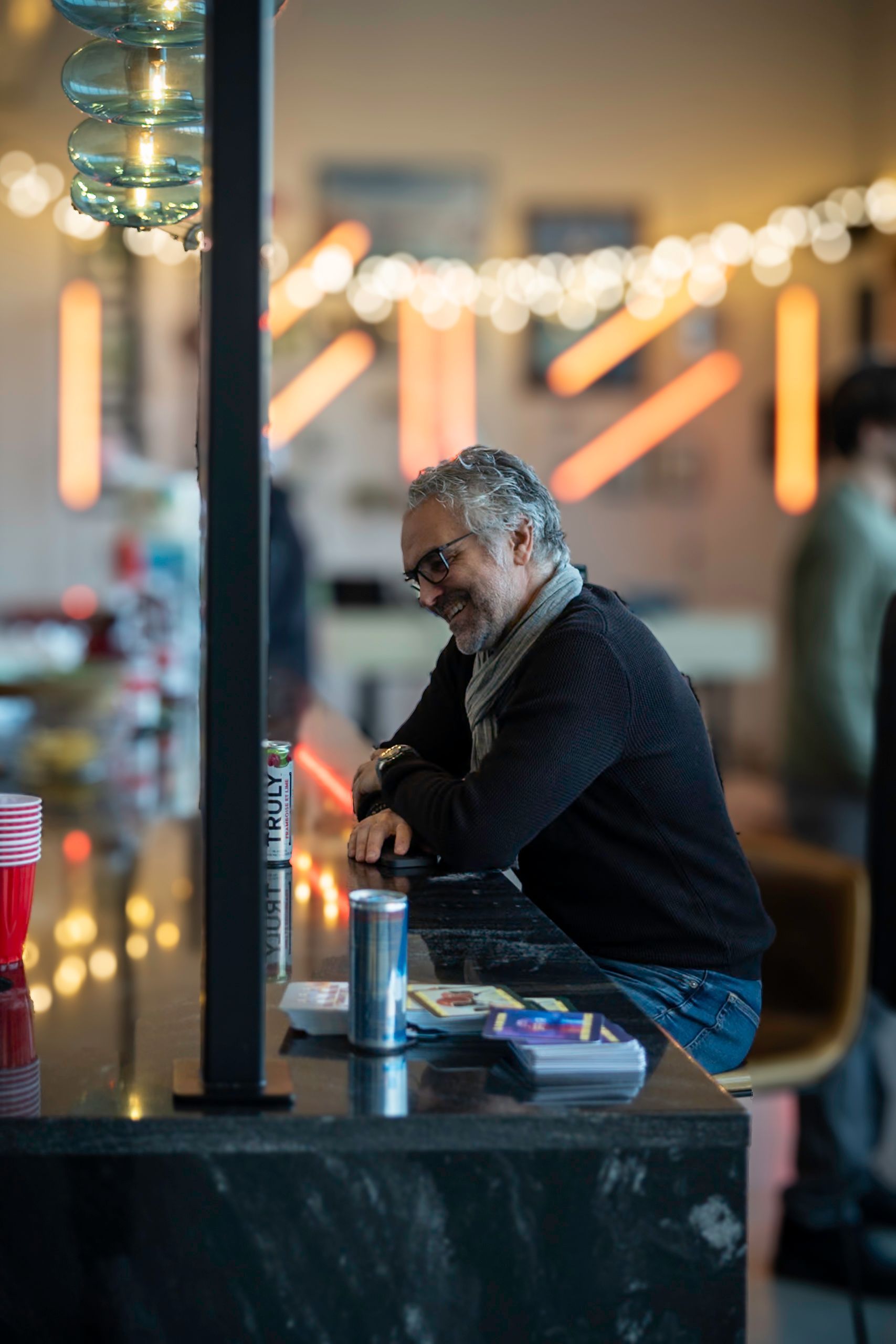 Man smiling, leaning on a bar, indoors. Modern setting with string lights and a can in front of him.