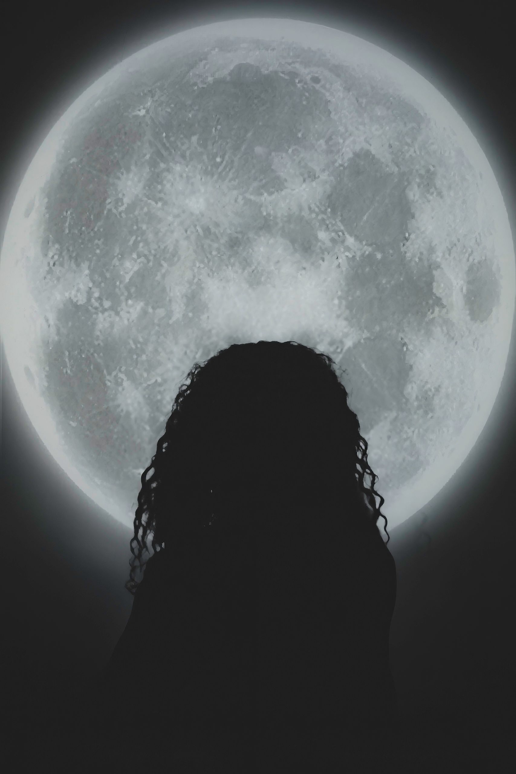 Silhouette of person with curly hair, in front of a large full moon in a dark night sky.