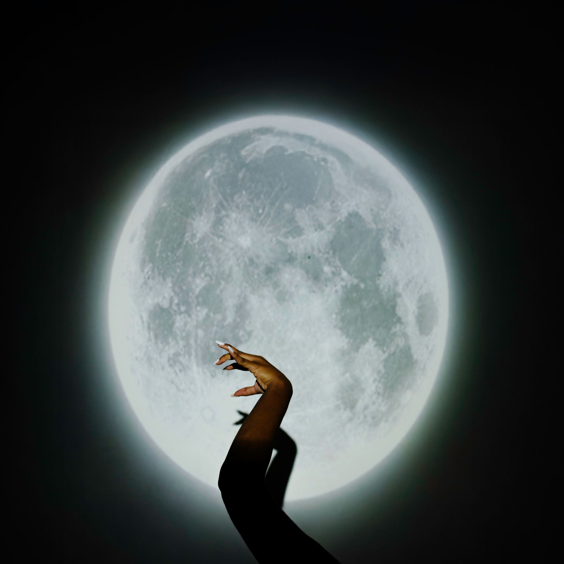 Hand with long nails reaching towards a large, bright moon against a dark background.