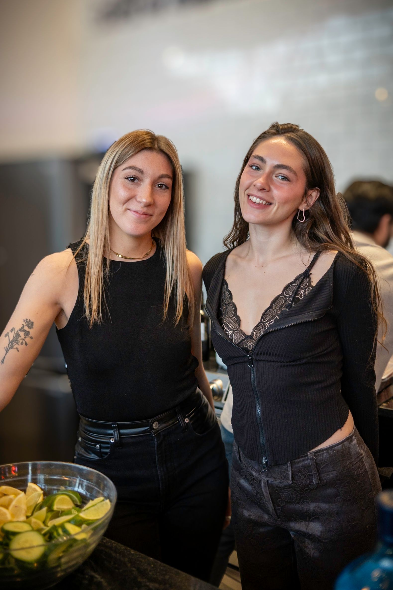 Two women smiling, one with blonde hair, the other with long dark hair, stand near a bowl of sliced cucumbers.