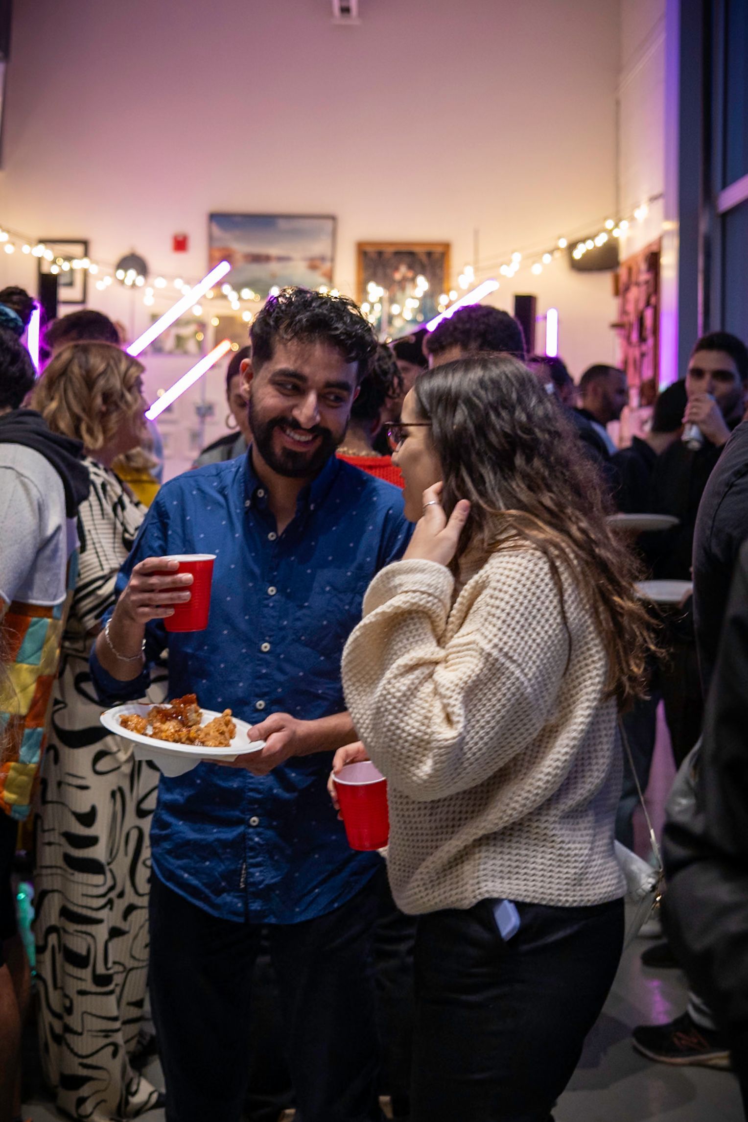 Man holding food smiles at woman with a red cup at a social gathering. String lights and artwork are in the background.