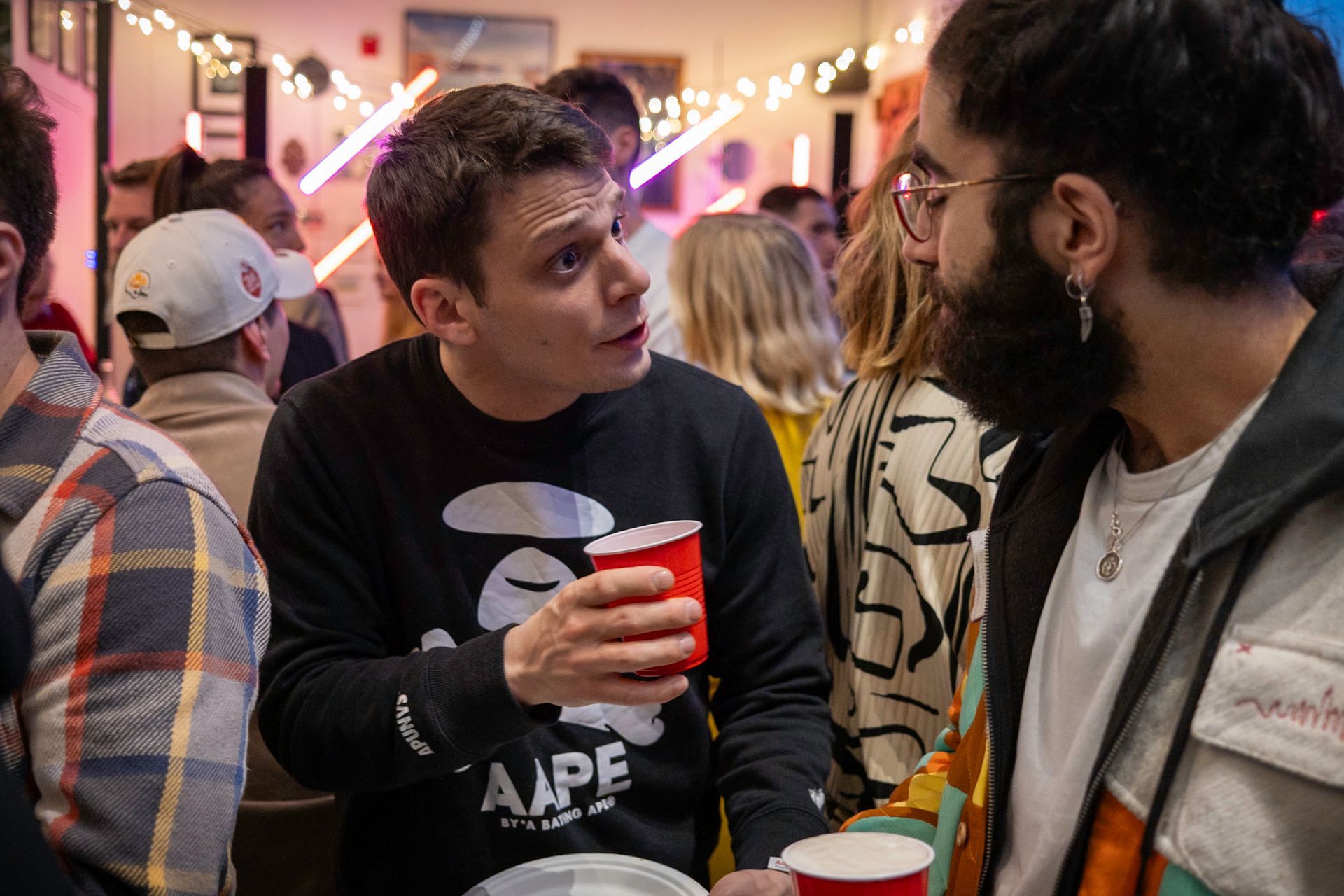 Man in black sweatshirt speaks to man with beard, both holding red cups, at a gathering. String lights in background.