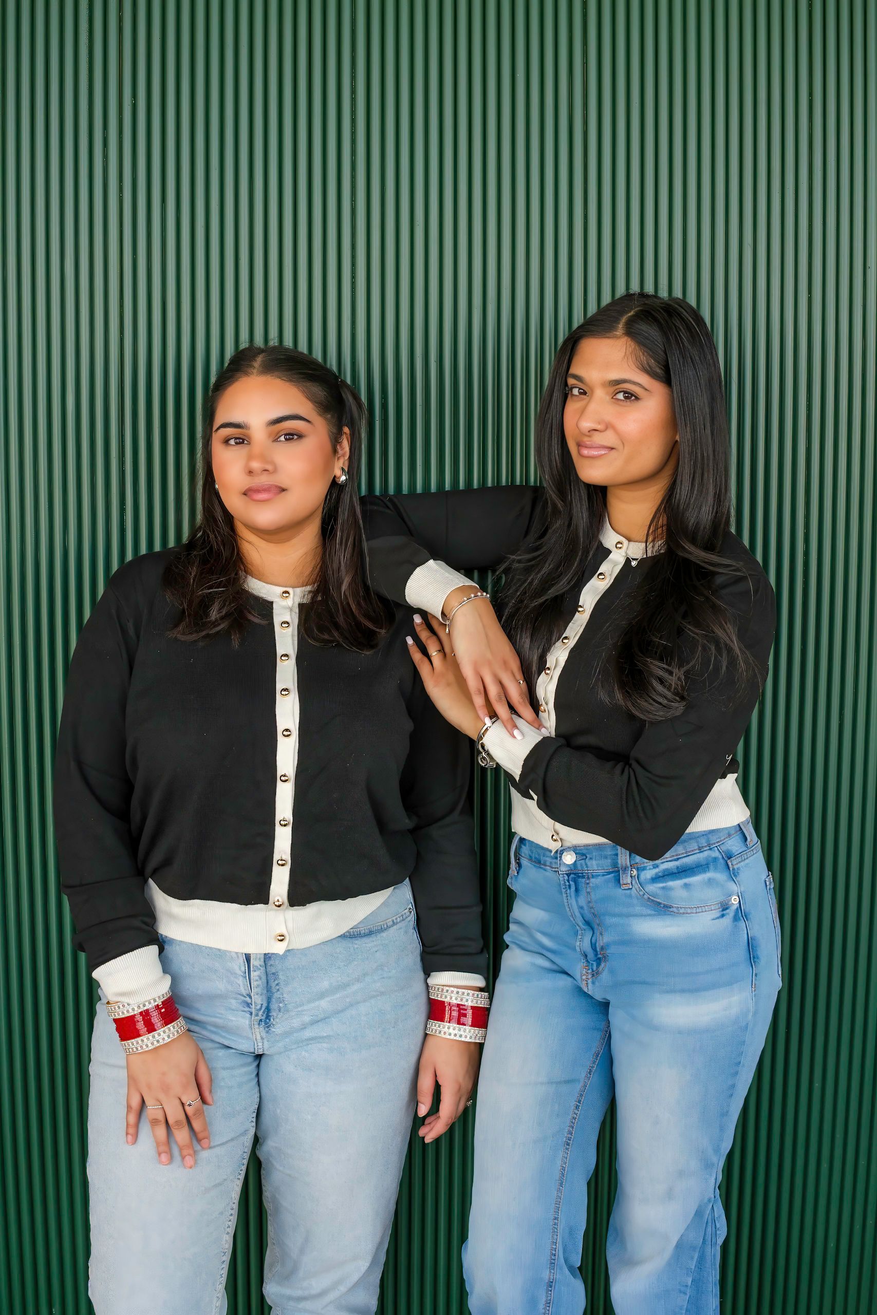 Two women wearing black sweaters with white trim and jeans lean against a green ribbed wall.