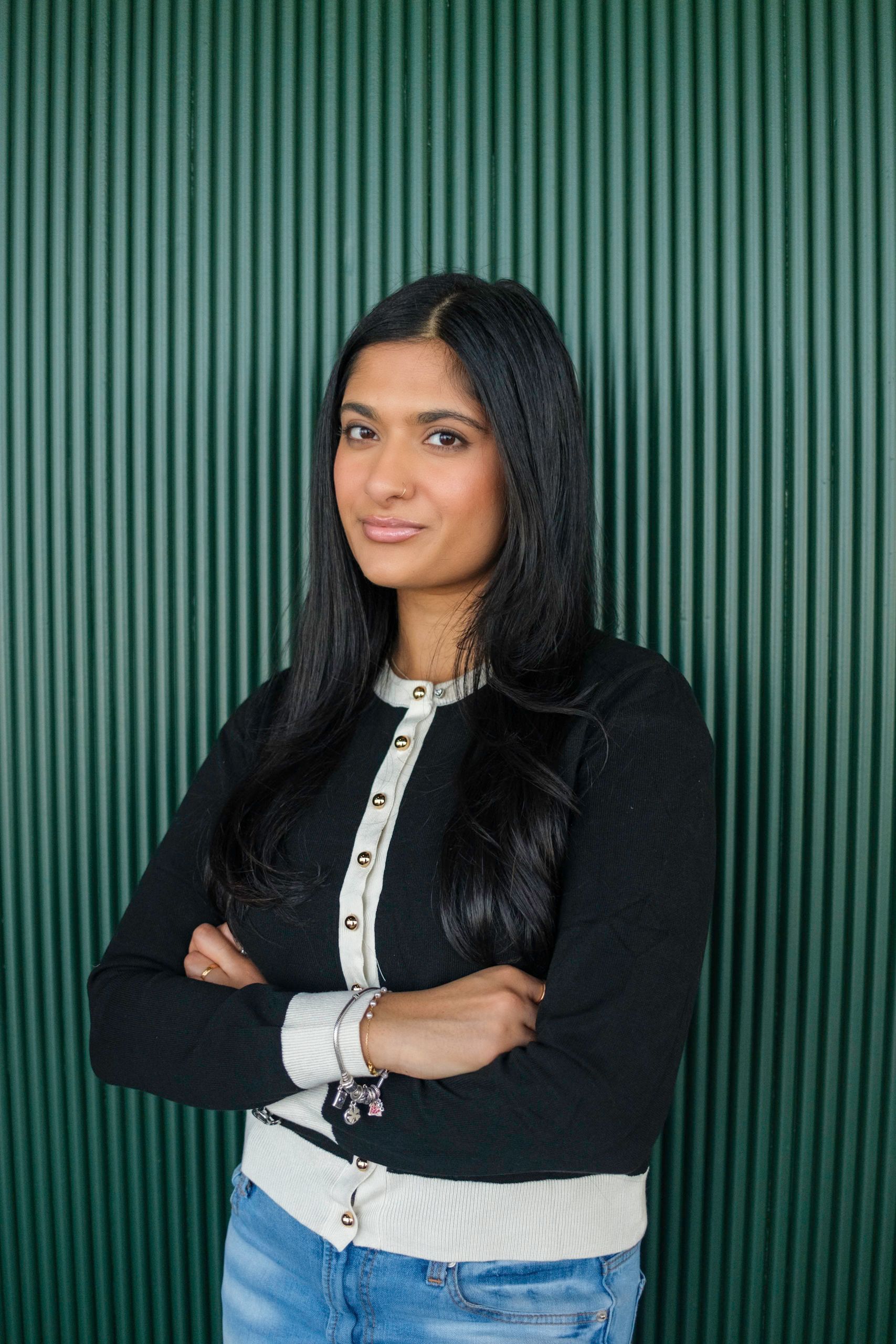 Woman with long dark hair, arms crossed, wearing a black cardigan and jeans, standing in front of a green wall.