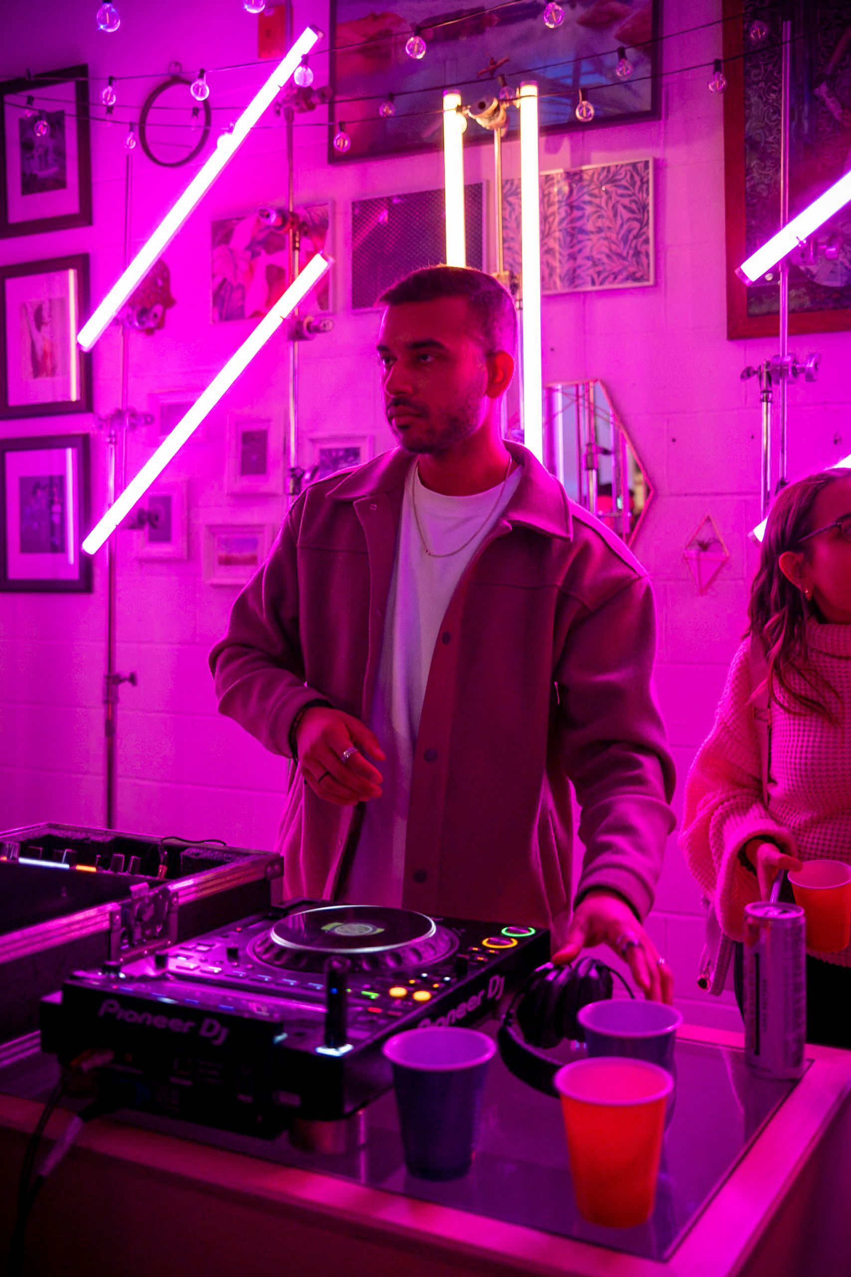 DJ at a party in a room with pink neon lights. He's operating turntables and mixers, with a few drinks in front of him.