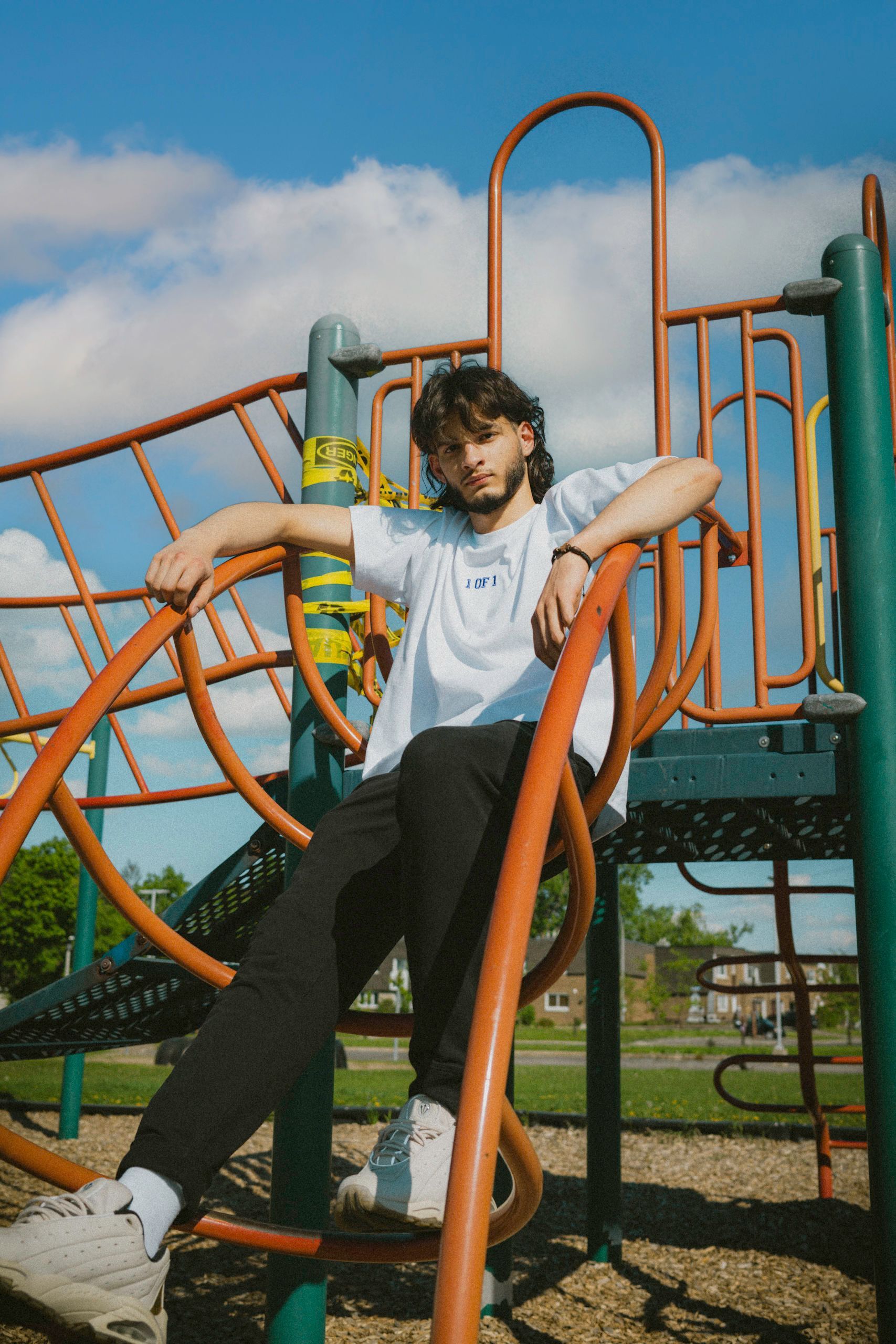 Man sitting on playground equipment, wearing a white shirt and black pants, with a blue sky background.