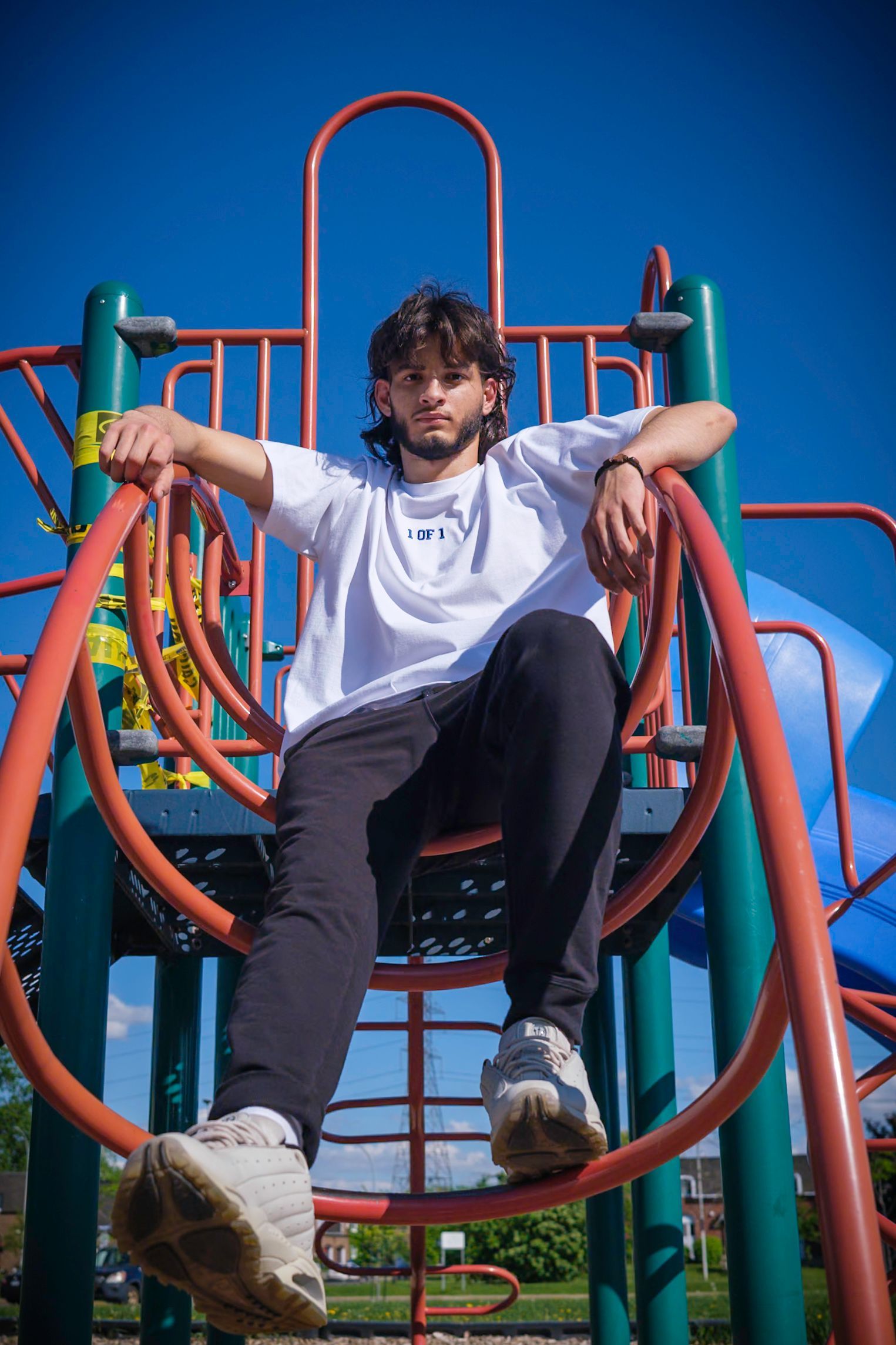 Man sitting on playground equipment, wearing a white t-shirt and black pants, against a blue sky.