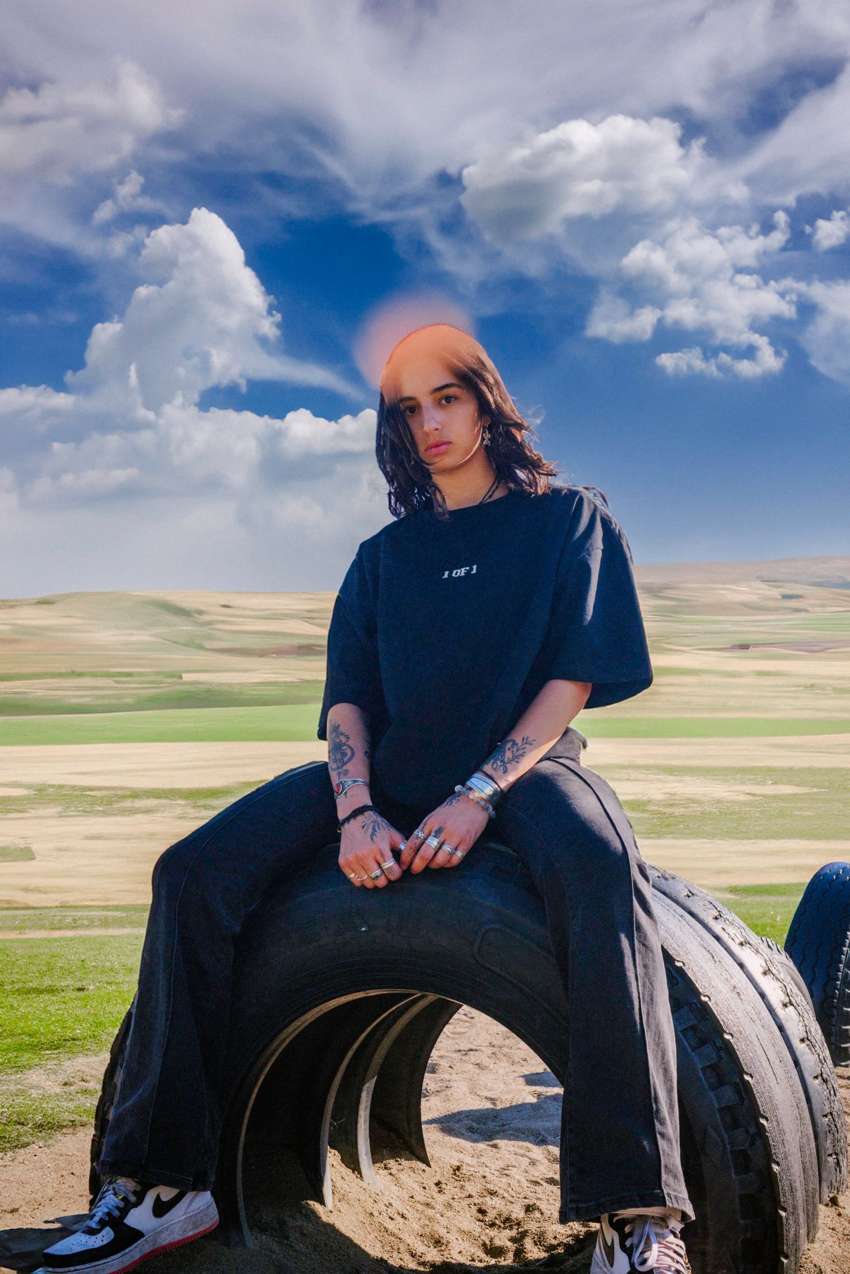 Person sitting on a tire, wearing black clothing, in a field with a cloudy sky.