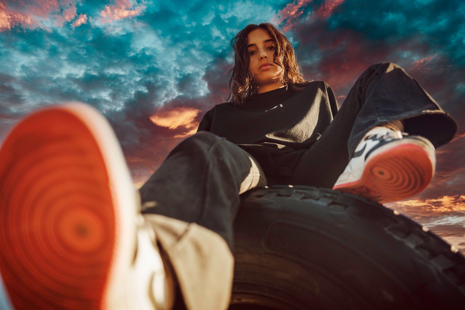 Person seated on a tire, feet extended towards the viewer, against a colorful sunset.