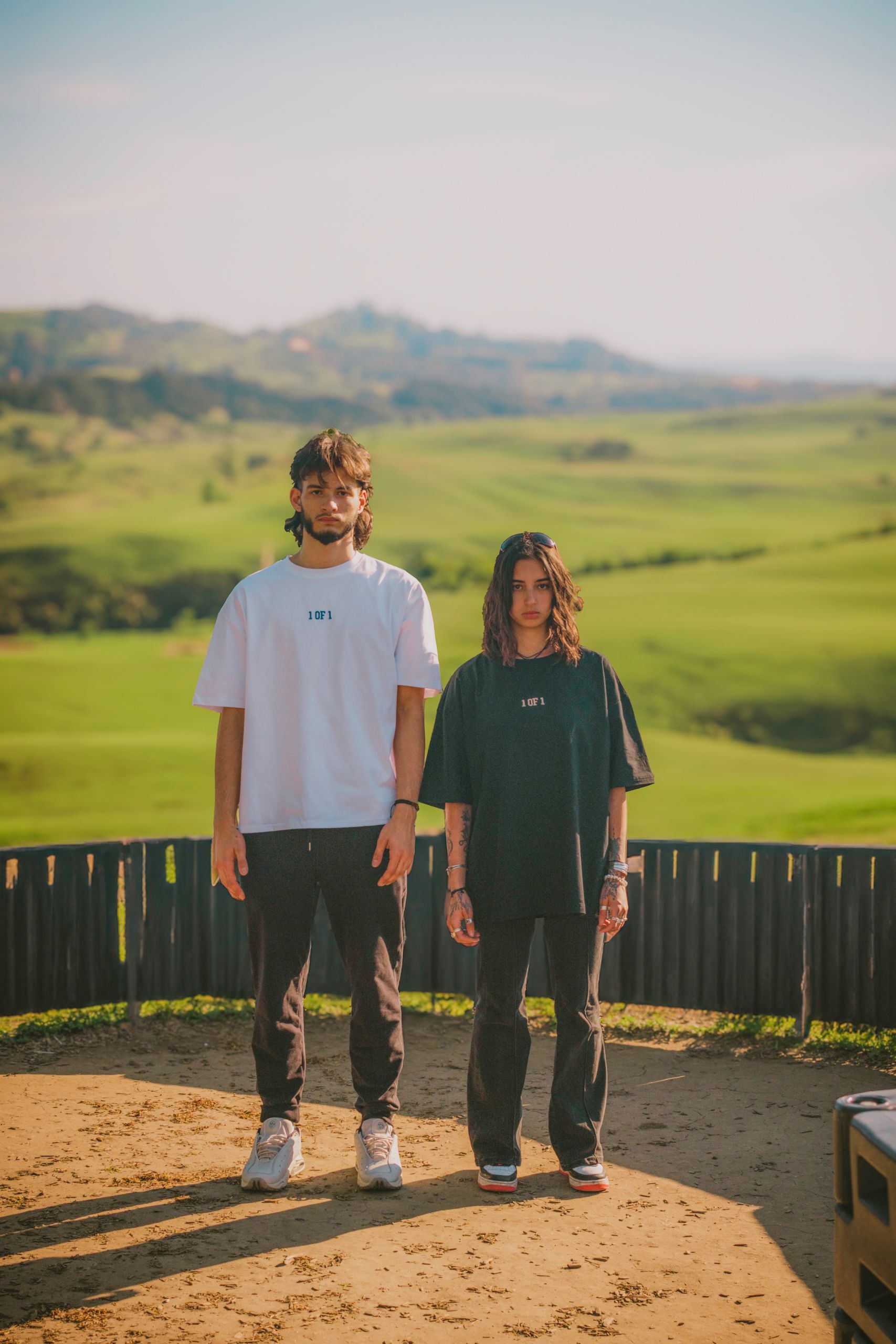 Two people stand together, wearing t-shirts and pants, in front of a green landscape.