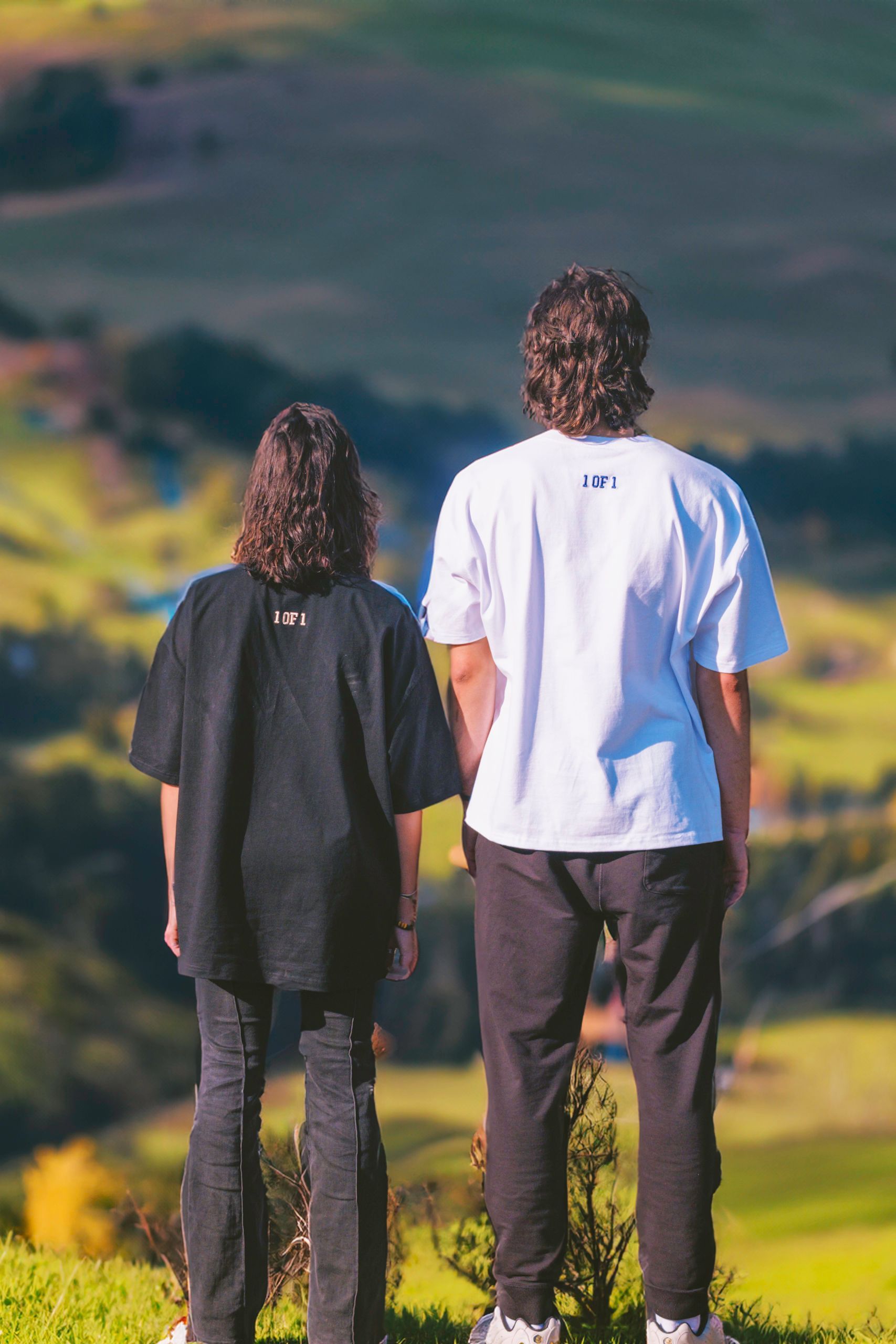 Two people stand on a hill overlooking a valley; one in black, the other in white, both wearing t-shirts.