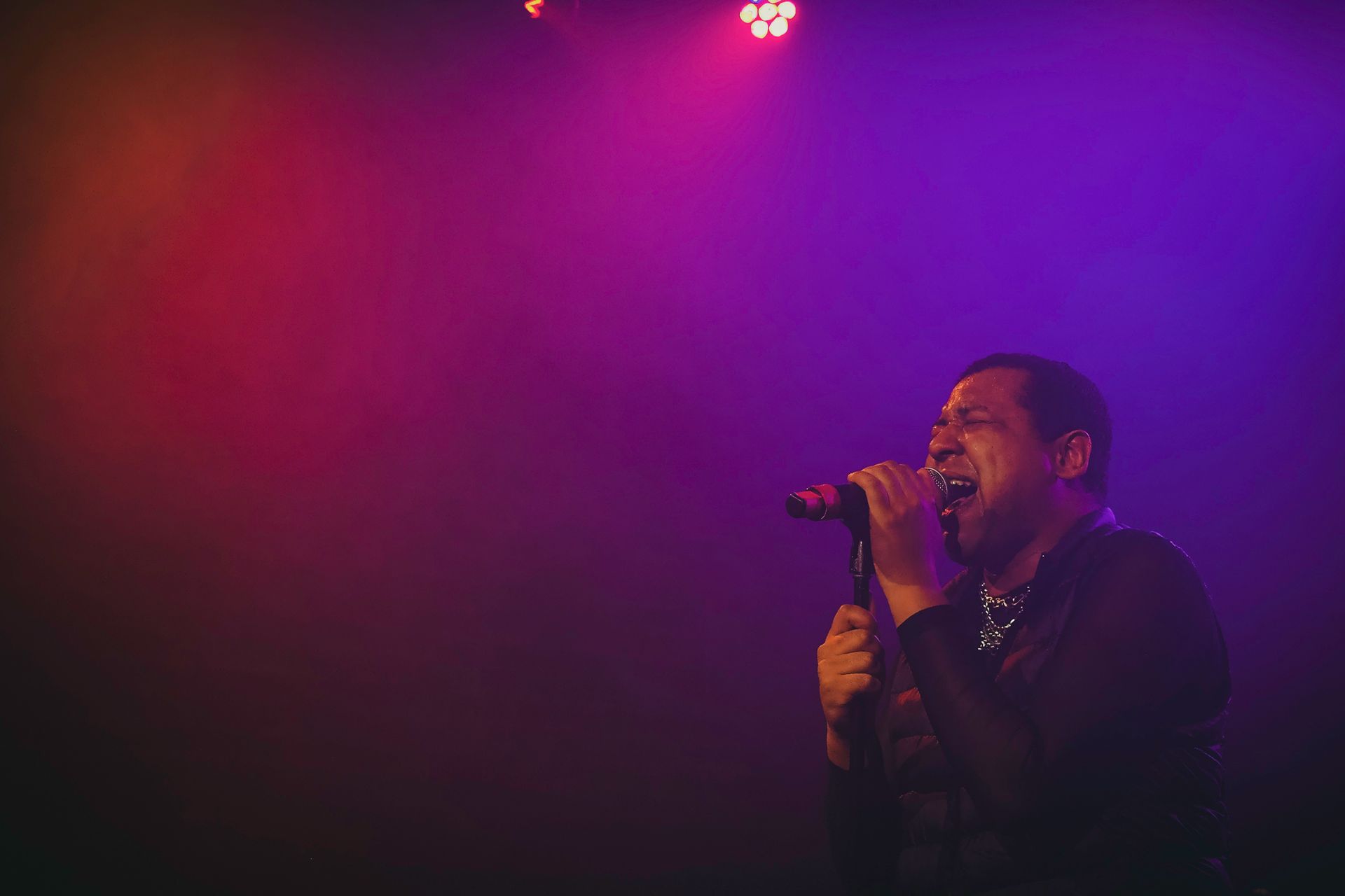 Singer performing on stage, lit by purple and red lights, holding a microphone, singing with an open mouth.