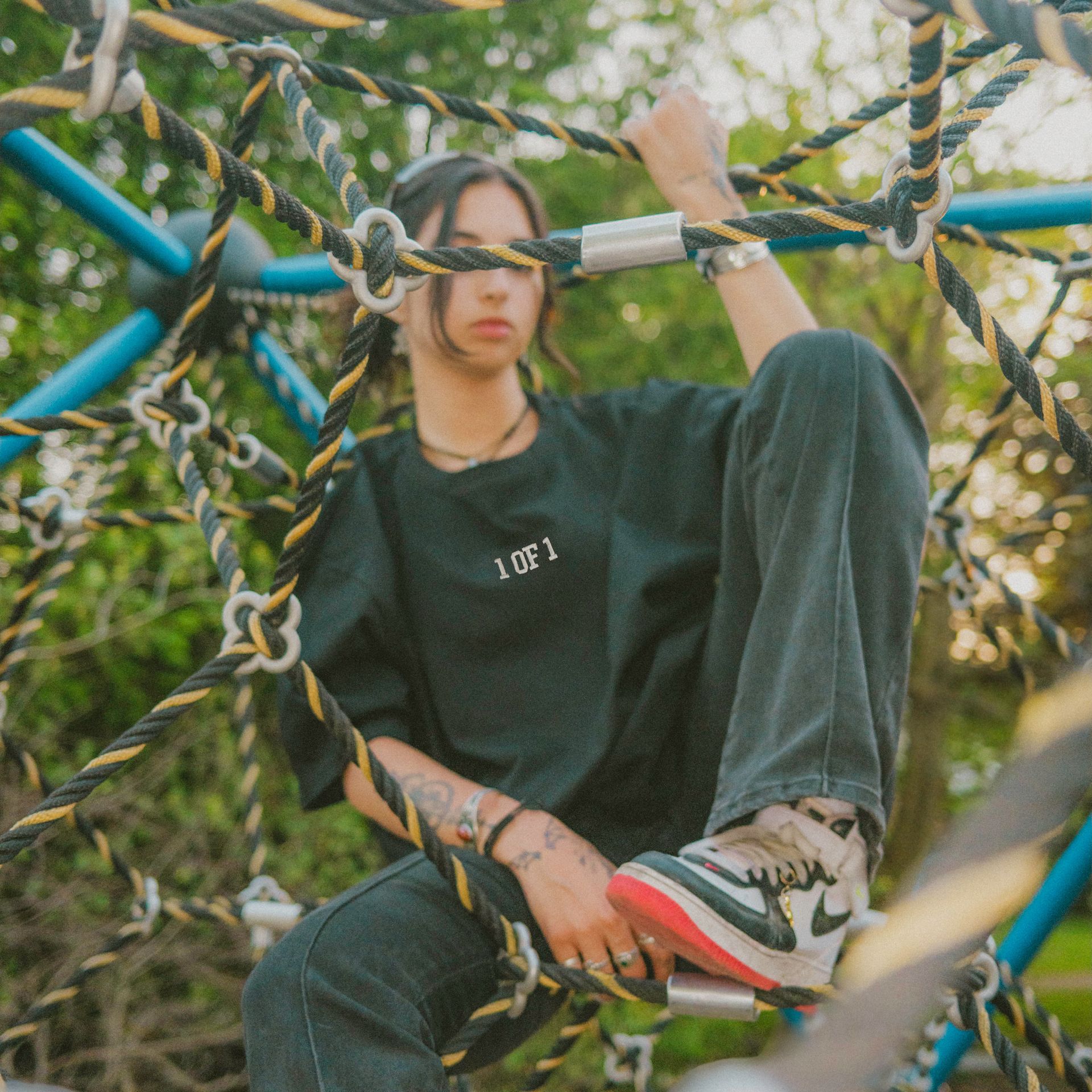 Person in black t-shirt and jeans sits on a climbing structure, holding onto ropes, wearing sneakers.
