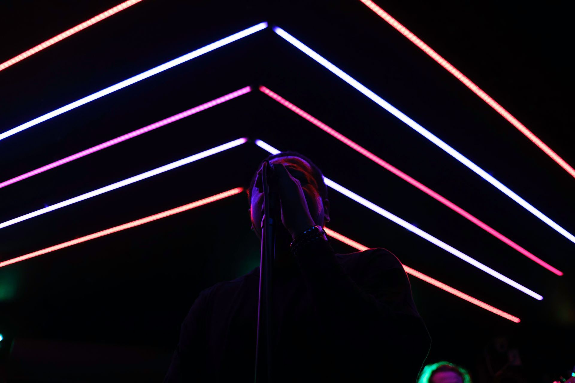Person singing into microphone, silhouetted against neon pink and white lines in a dark venue.