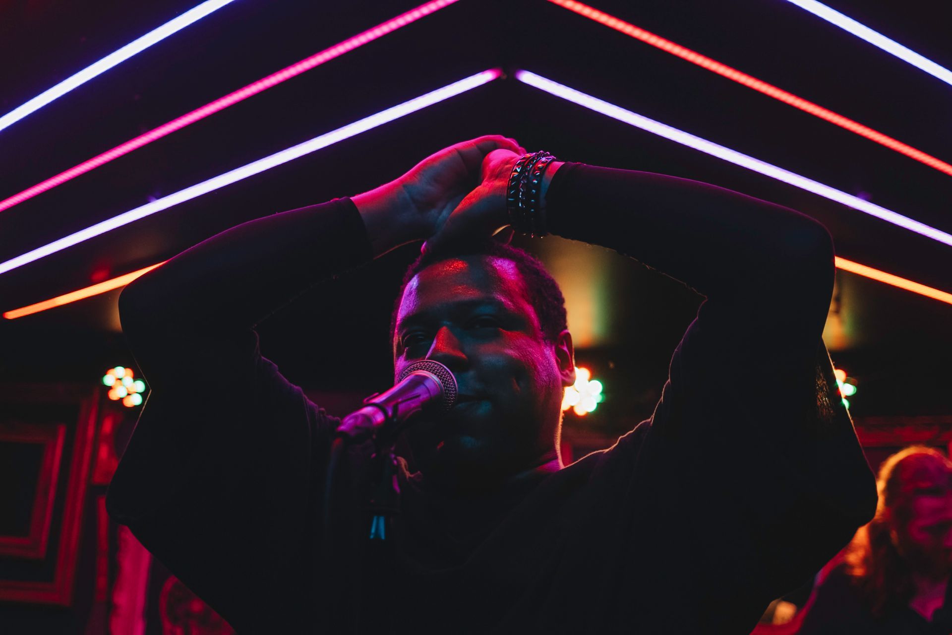 Man singing into a microphone, arms raised. Pink and purple neon lights in background.