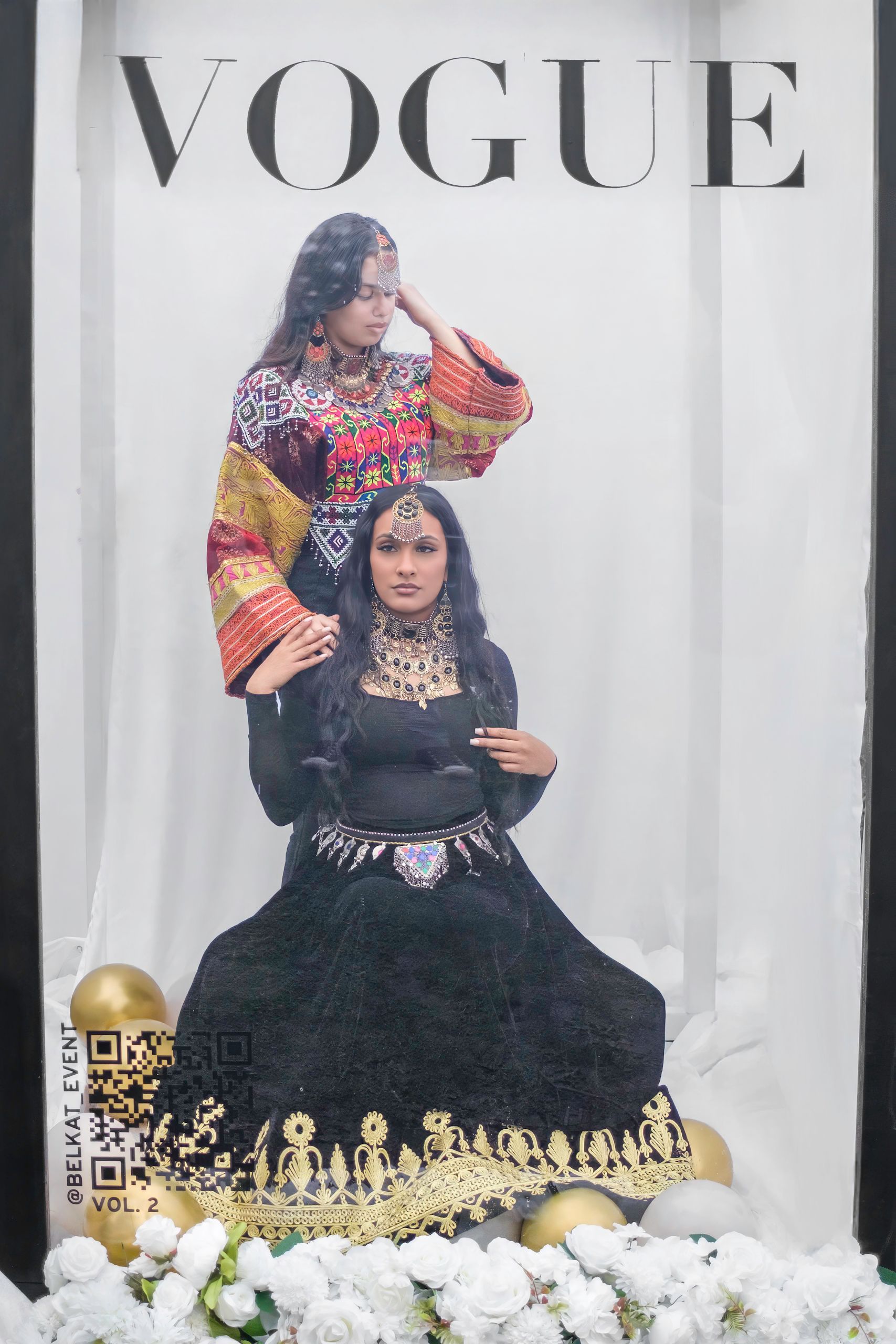 Two women pose in traditional clothing, framed by a Vogue cover. Gold and white floral decorations at the base.