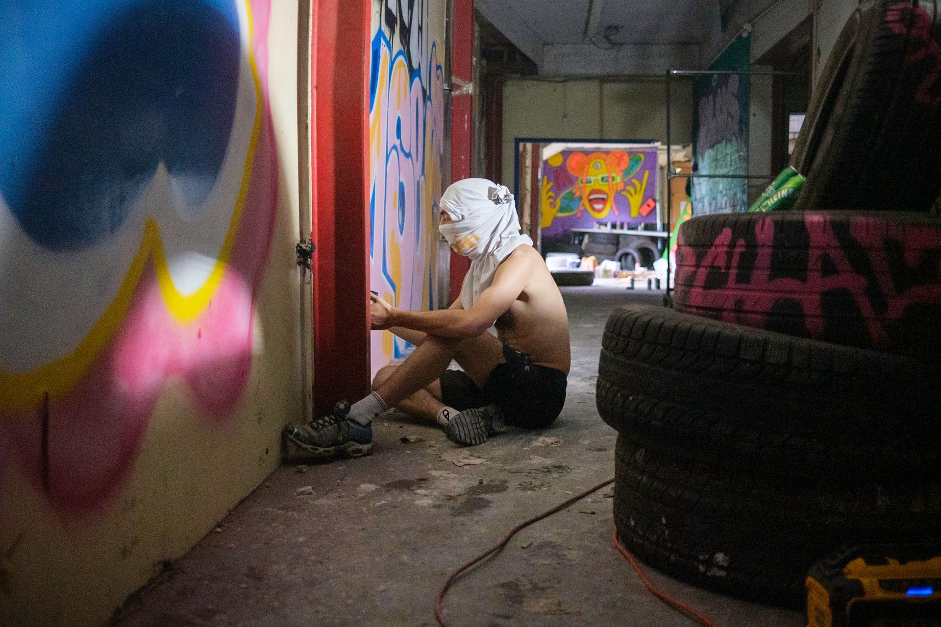 Person spray-painting graffiti in a dimly lit, cluttered space. They wear a face covering and sit near stacked tires.