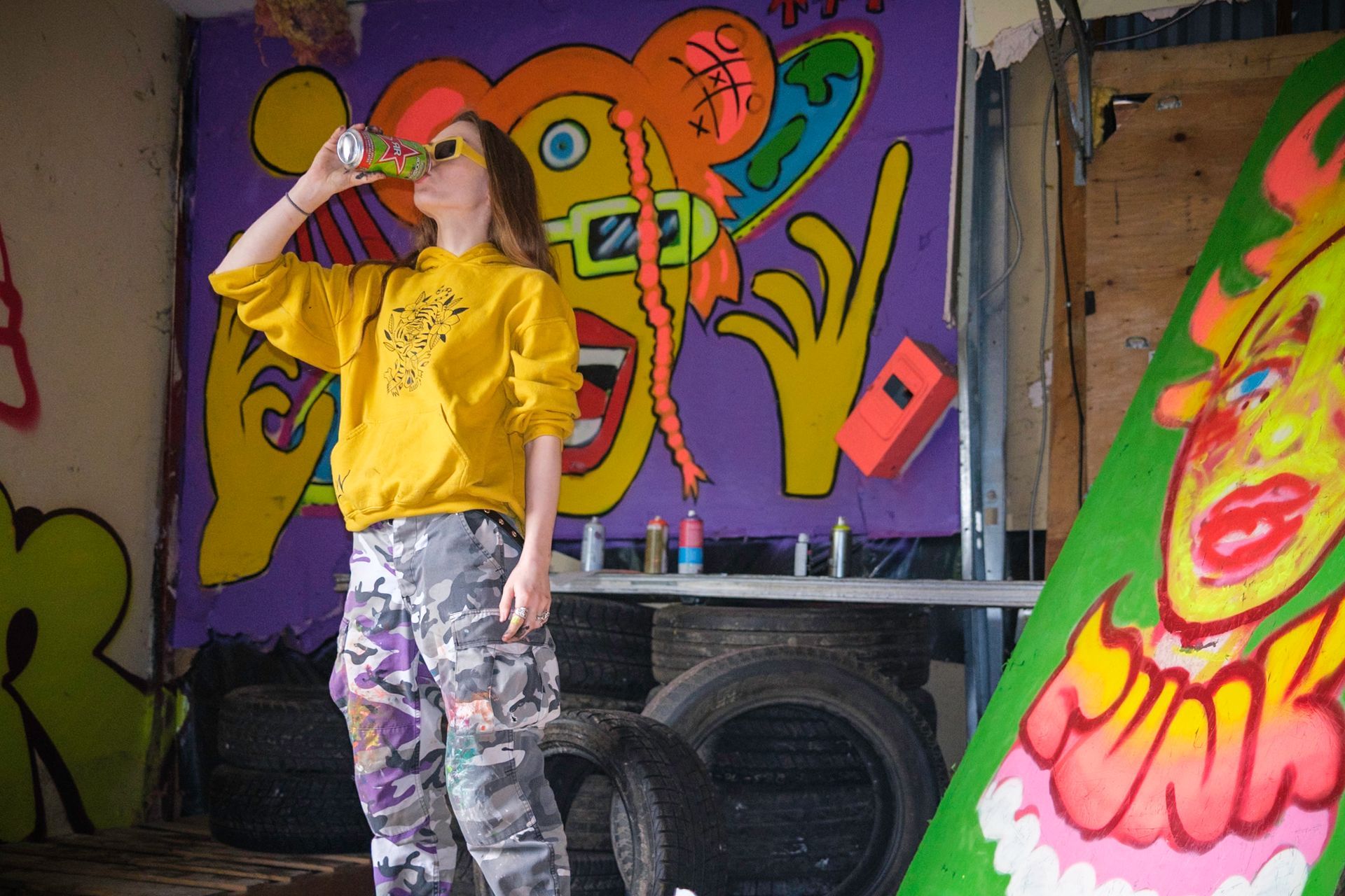 Person in yellow hoodie drinks from a can in front of graffiti art; indoor setting with tires and artwork.