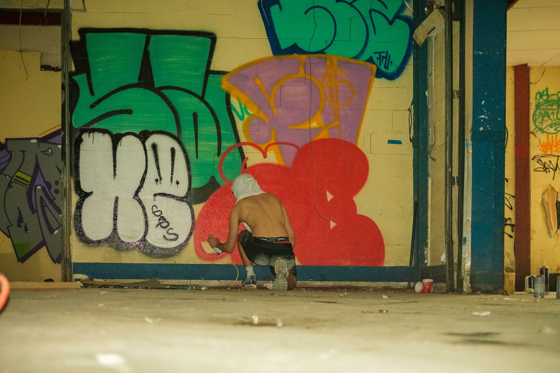 Person spray-painting graffiti in a dimly lit, abandoned building. Colorful tags on the wall.