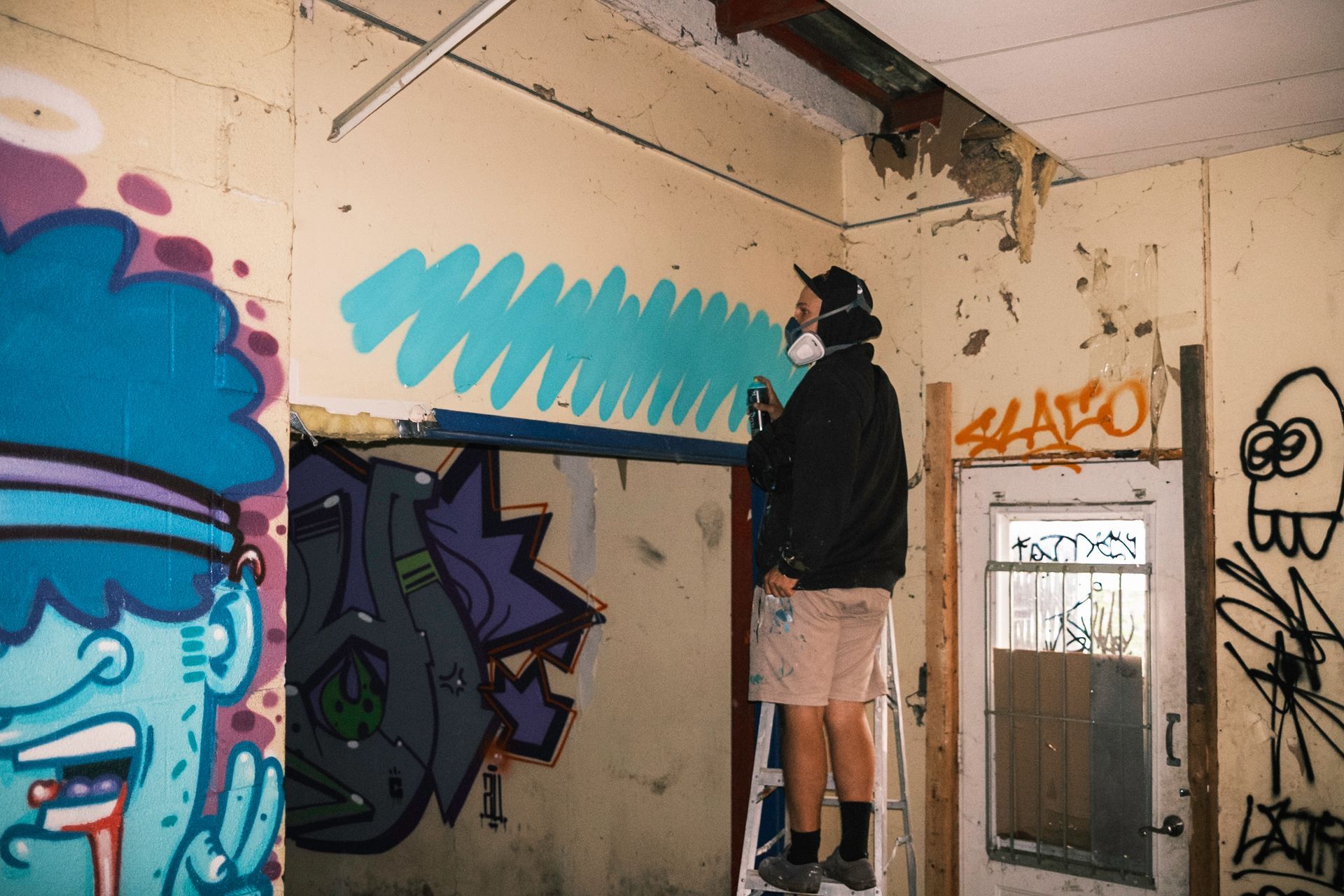 Person on ladder painting graffiti in a dilapidated room with existing artwork. Blue and teal paint.