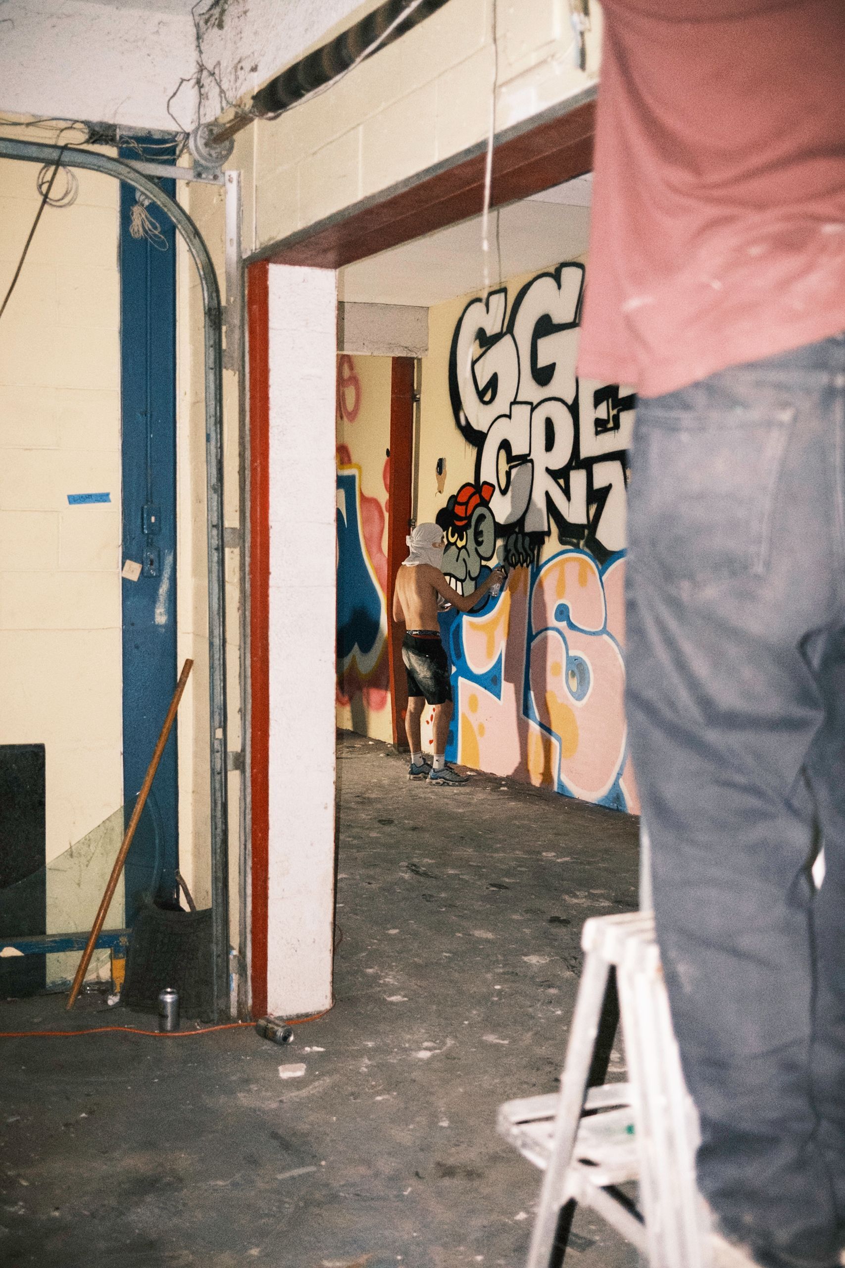 Person paints graffiti inside building; red, white, blue colors. Another person on stool in the foreground.