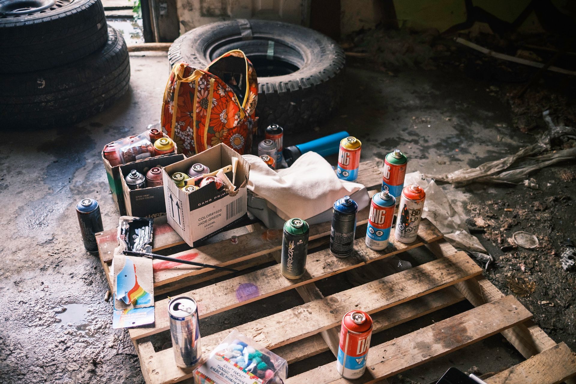 A wooden pallet with spray paint cans, an open box with art supplies, and a bag in a grungy garage.
