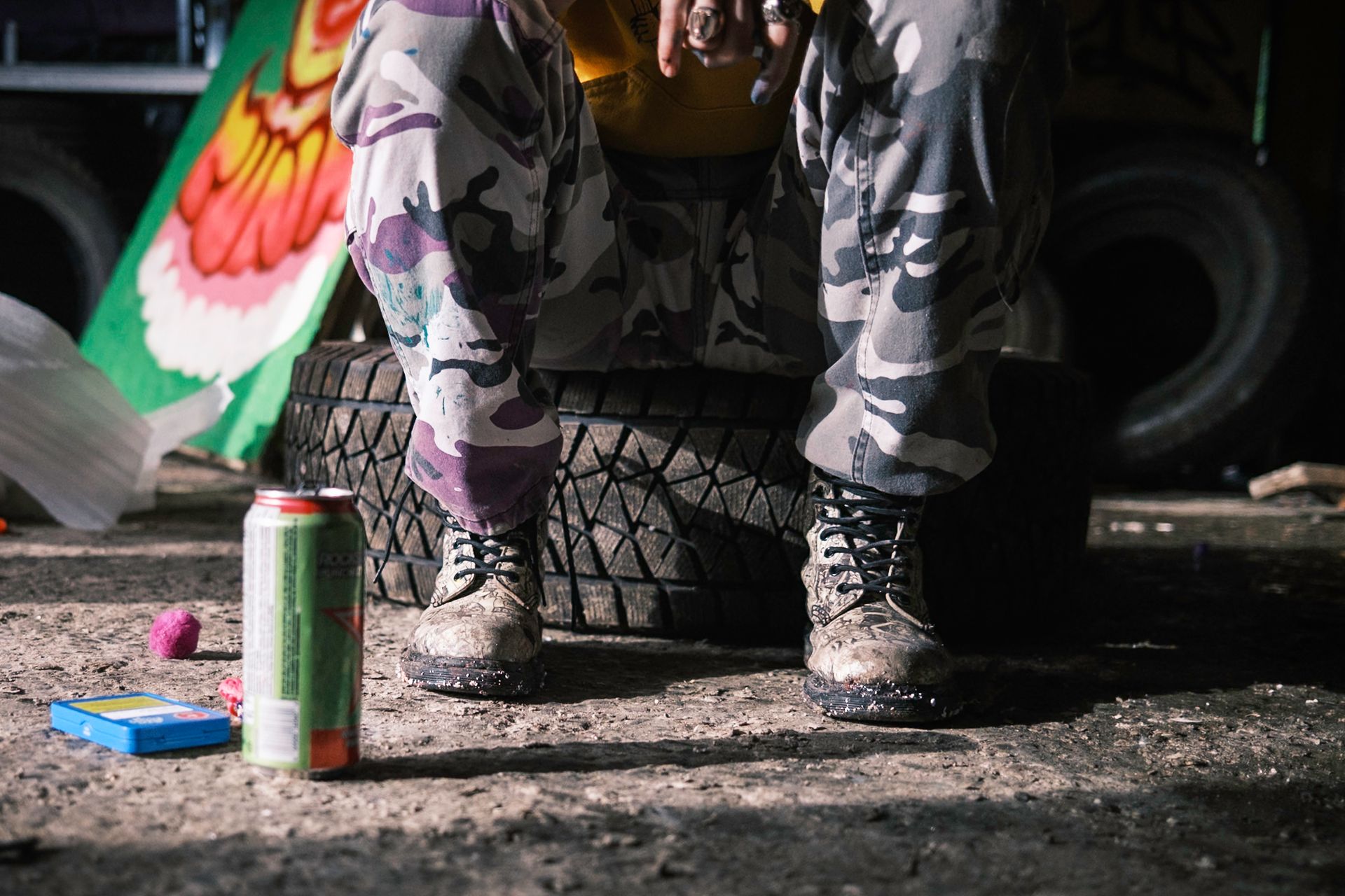 Person in camouflage pants and boots sits on a tire, surrounded by debris. A can and toy are on the ground.