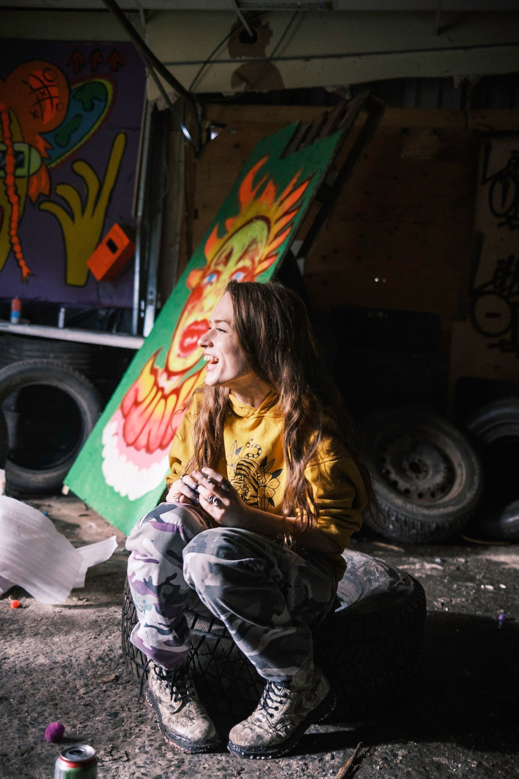Woman in yellow hoodie and camo pants squats, smiling, in front of colorful artwork and tires.