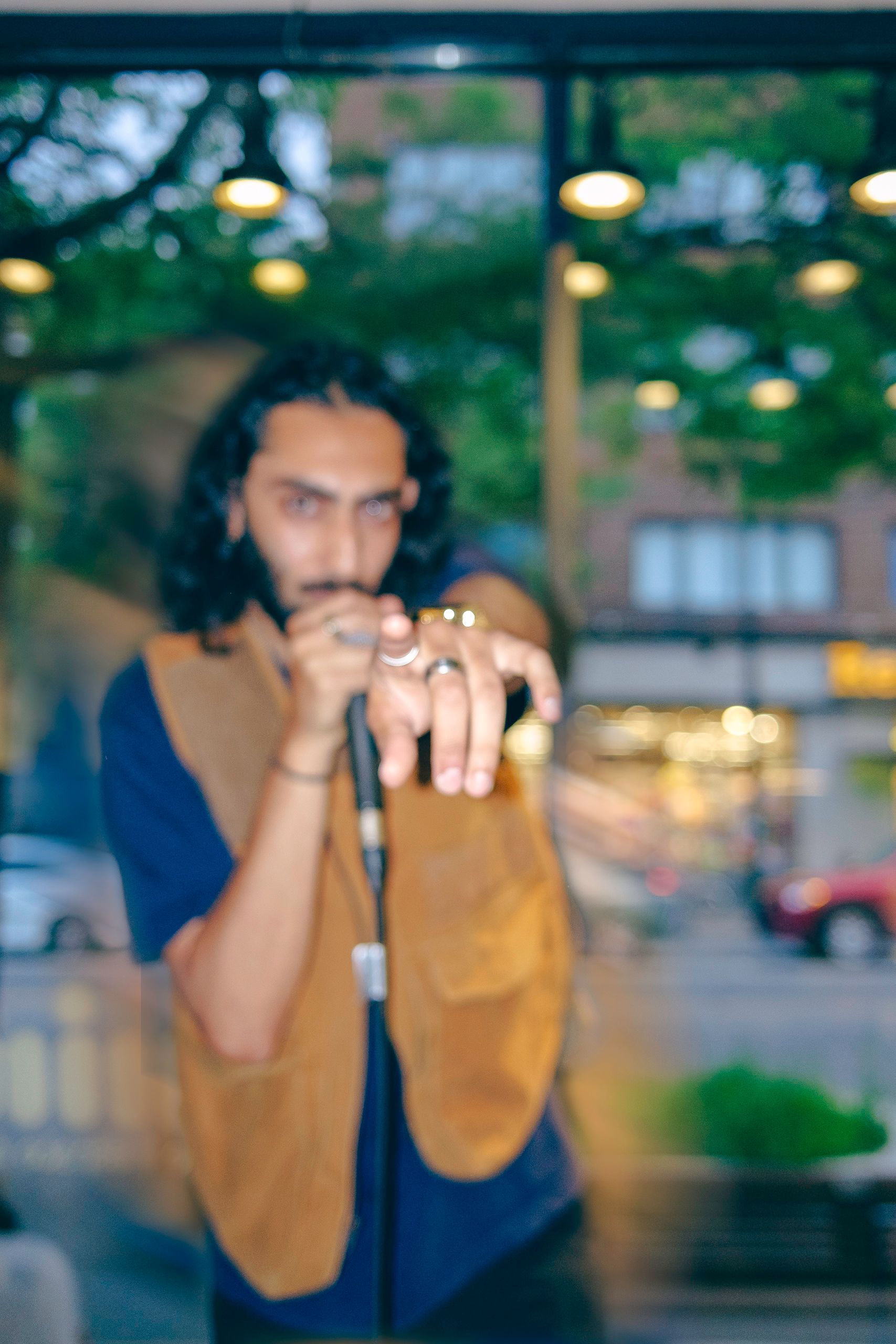 Man with long hair and microphone, wearing a vest, pointing forward. Blurred background of a street.