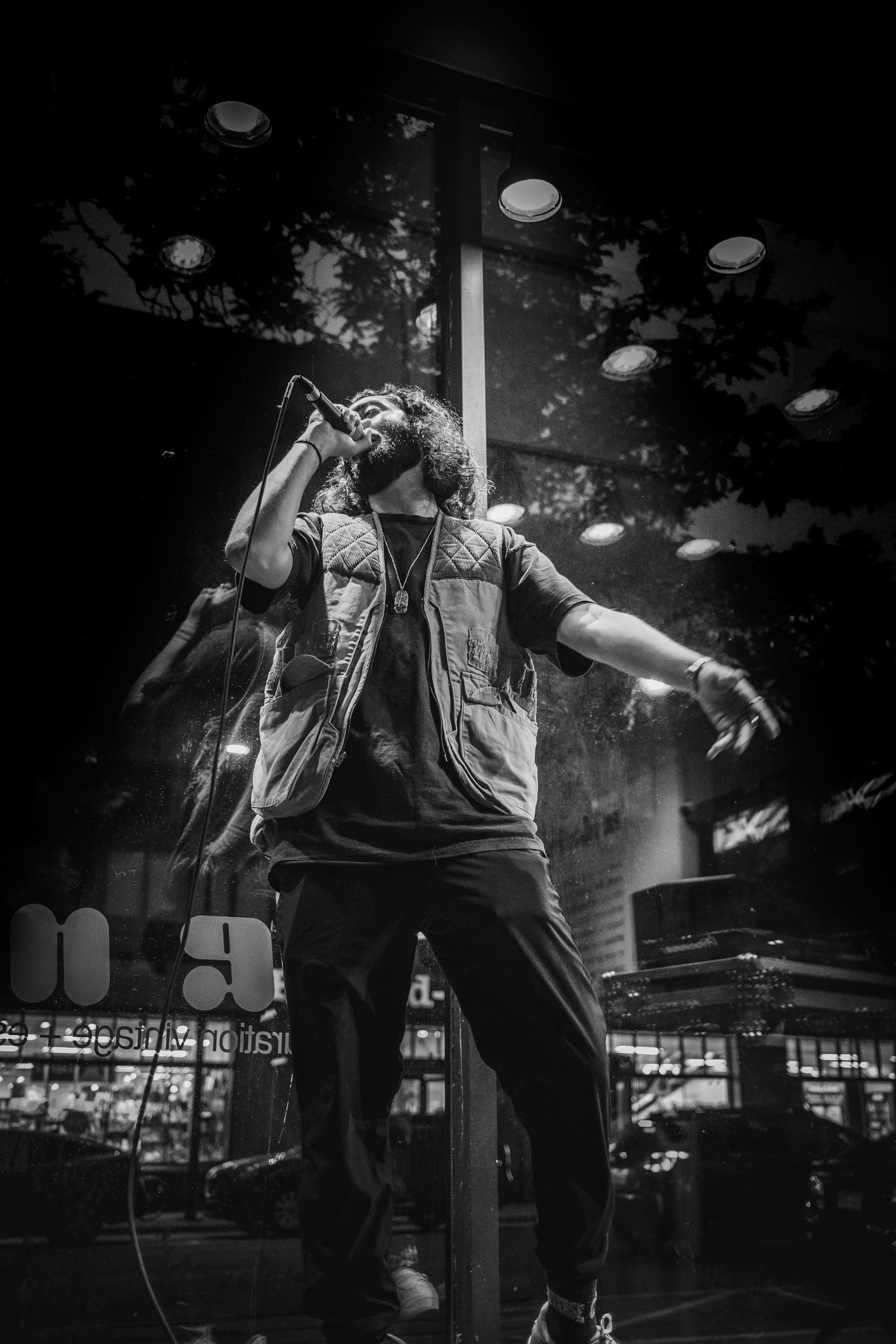 A performer sings into a microphone, wearing a vest and dark pants. Backlit window in the background.