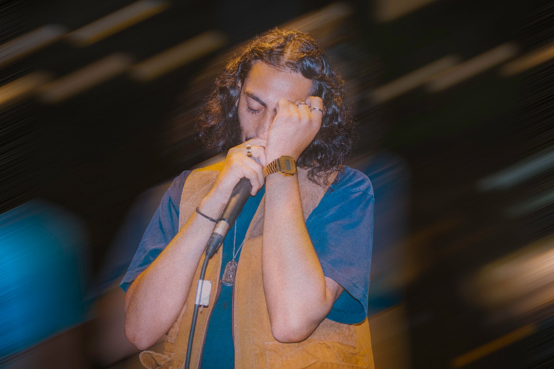 Man singing into a microphone, covering his eyes with his hand. He wears a blue shirt and brown vest on a stage.
